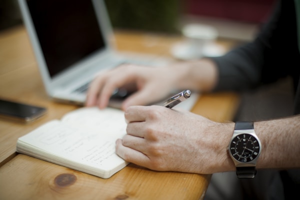 A man writing on a notebook while using a laptop.