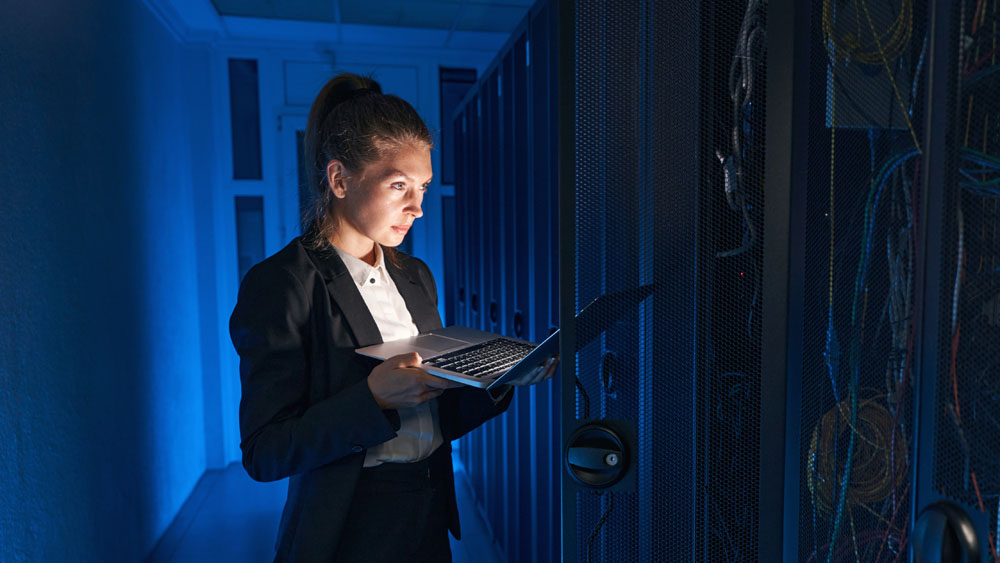 A woman standing in front of a server holding a laptop.