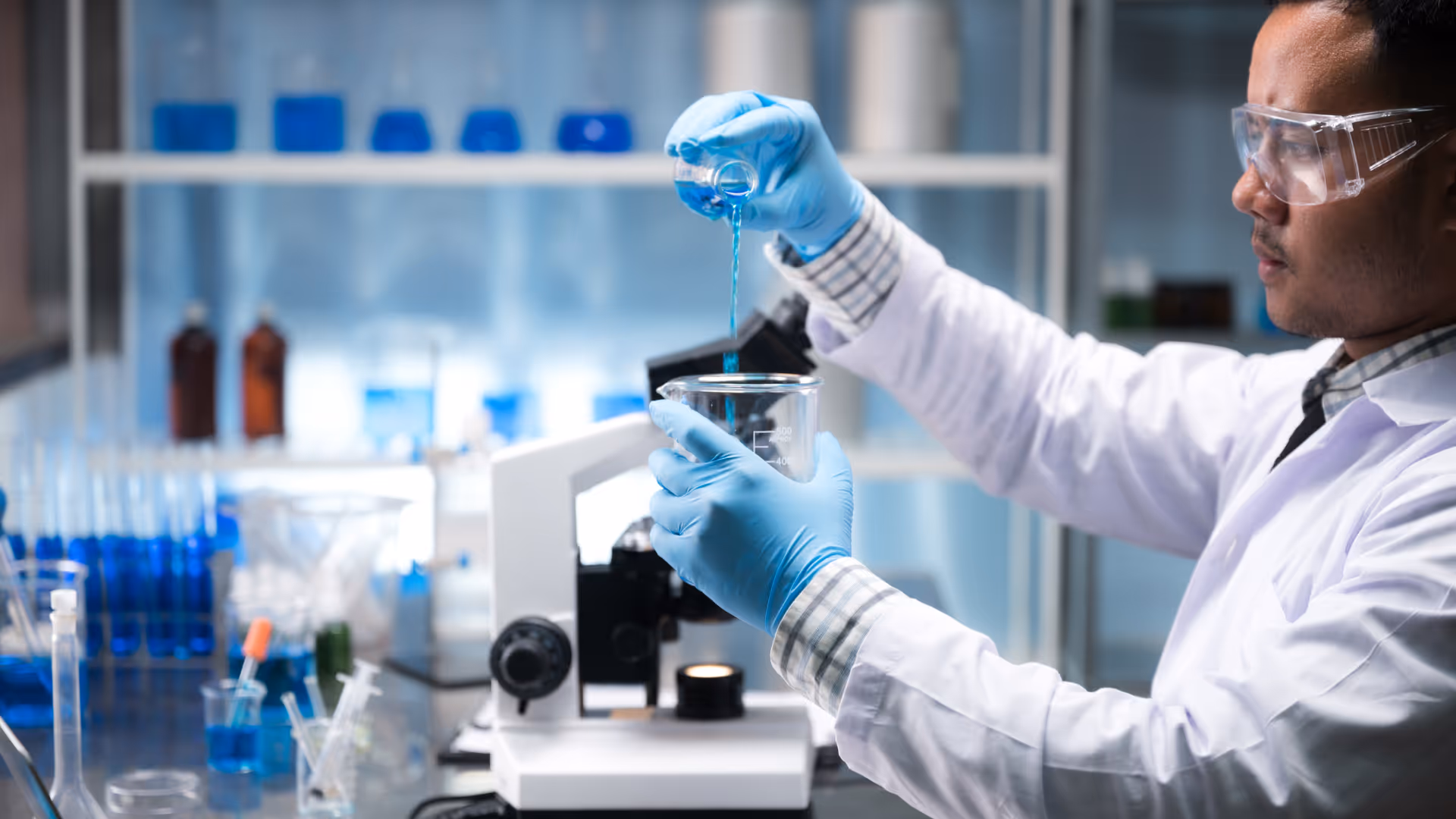 Scientist wearing gloves and goggles pouring blue liquid from a small beaker into a larger beaker in a laboratory.