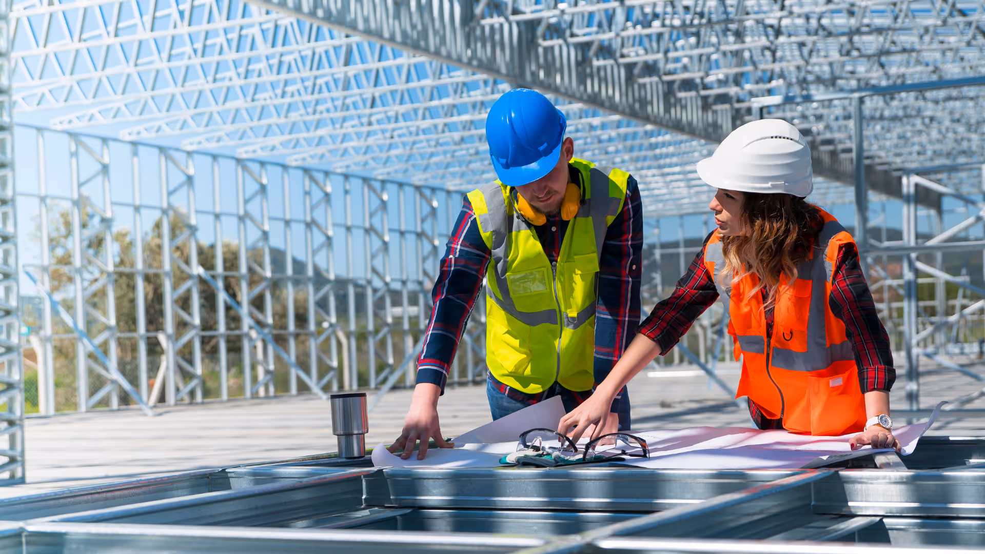 Two construction workers wearing safety vests and helmets reviewing blueprints inside a steel framework building under construction.