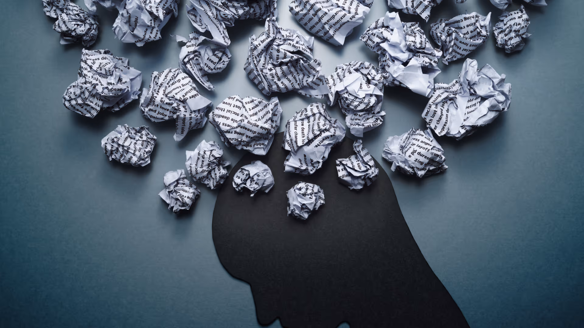 Silhouette of a human head with crumpled pieces of printed paper emerging from it against a gray background.