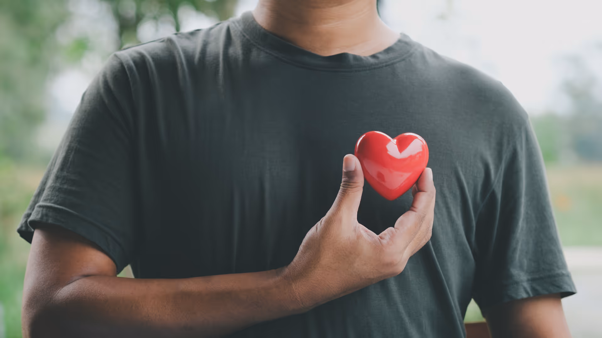 Person wearing a dark shirt holding a small red heart close to their chest.