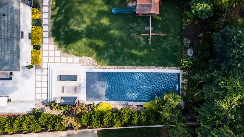 An overhead aerial view of a luxury rectangular swimming pool featuring an integrated tanning ledge, custom stone paver patio decking, and lush green landscaping borders in a Raleigh, North Carolina backyard.