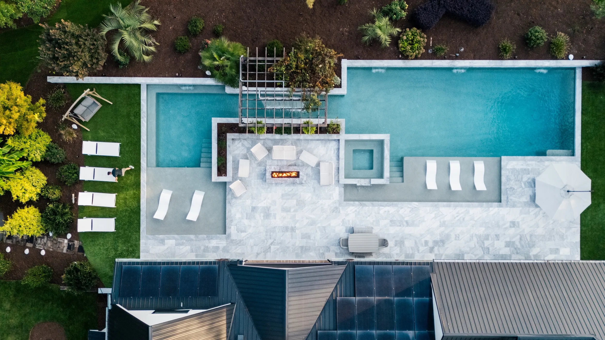 A top-down aerial view of a luxury rectangular swimming pool with an integrated spa, custom stone patio decking, a modern built-in outdoor fire pit with lounge seating, and surrounding landscaping by a Raleigh, NC pool builder.