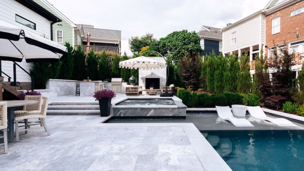 A luxury geometric swimming pool with integrated lounge steps, surrounded by a modern light stone patio, private white cabana seating, and a lush green privacy tree line in a Raleigh, NC backyard.