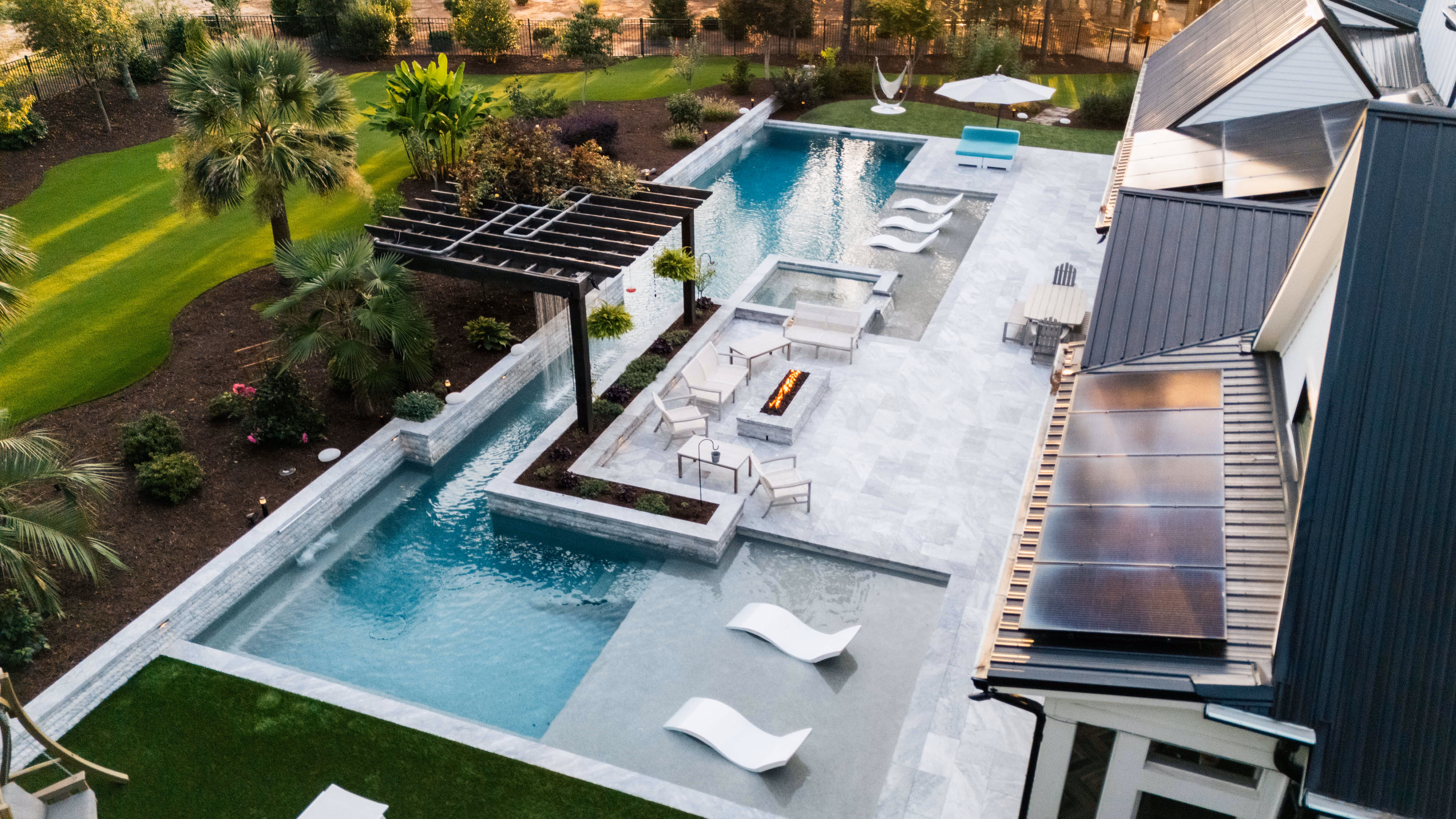 An illuminated luxury backyard retreat in Raleigh, NC, featuring a custom inground swimming pool, modern patio lounge, and professional landscape lighting by Currin Outdoor Living.