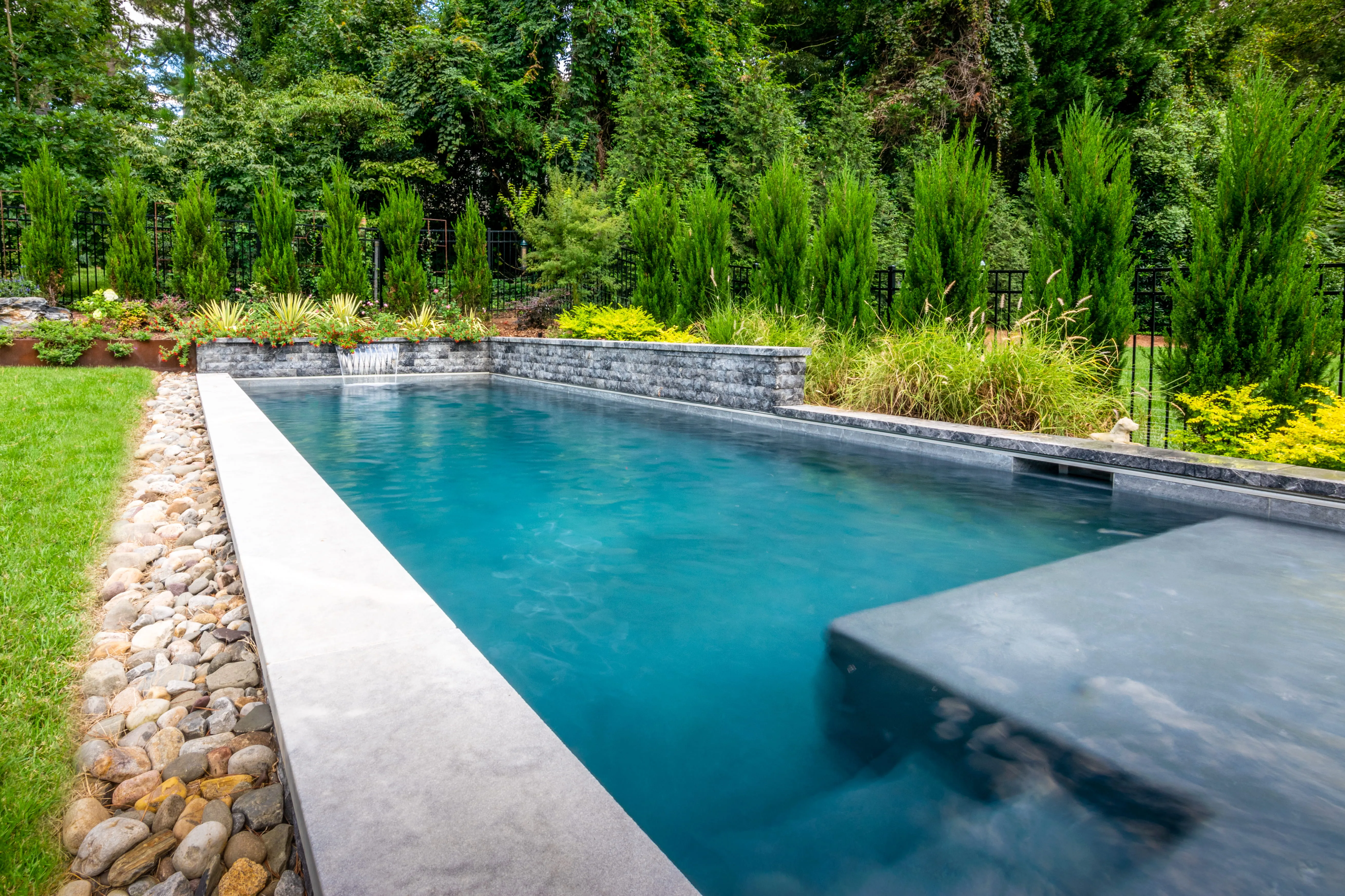 A striking, geometric custom inground pool by Currin Outdoor Living, featuring professional hardscape decking, modern perimeter lighting, and lush privacy landscaping in a Raleigh, NC backyard.