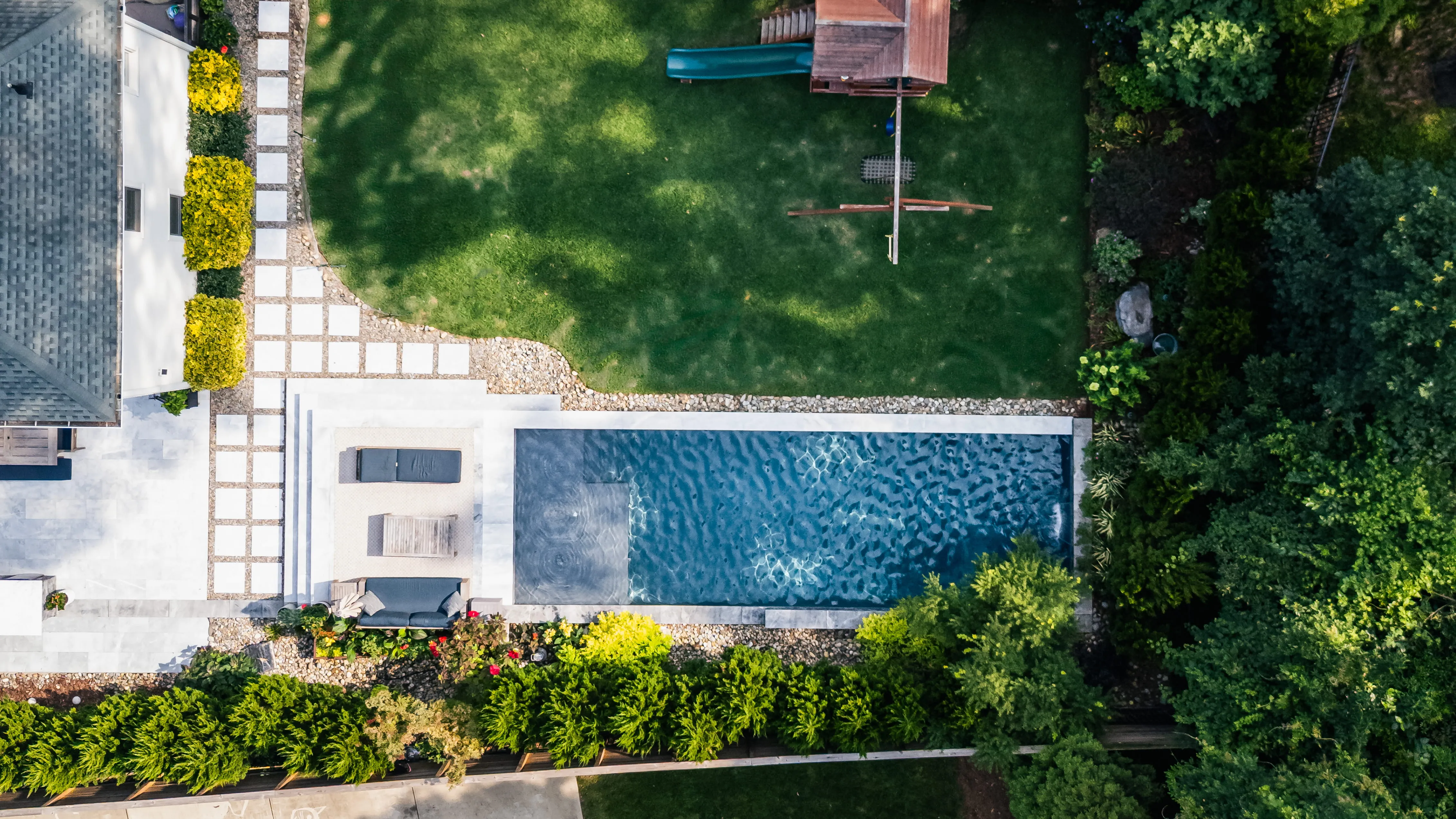 Top-down aerial view of a luxury custom rectangular inground swimming pool featuring a tanning ledge with in-pool lounge chairs, a sprawling modern stone paver patio, and professional backyard landscape design built by an outdoor living company in Raleigh, NC.