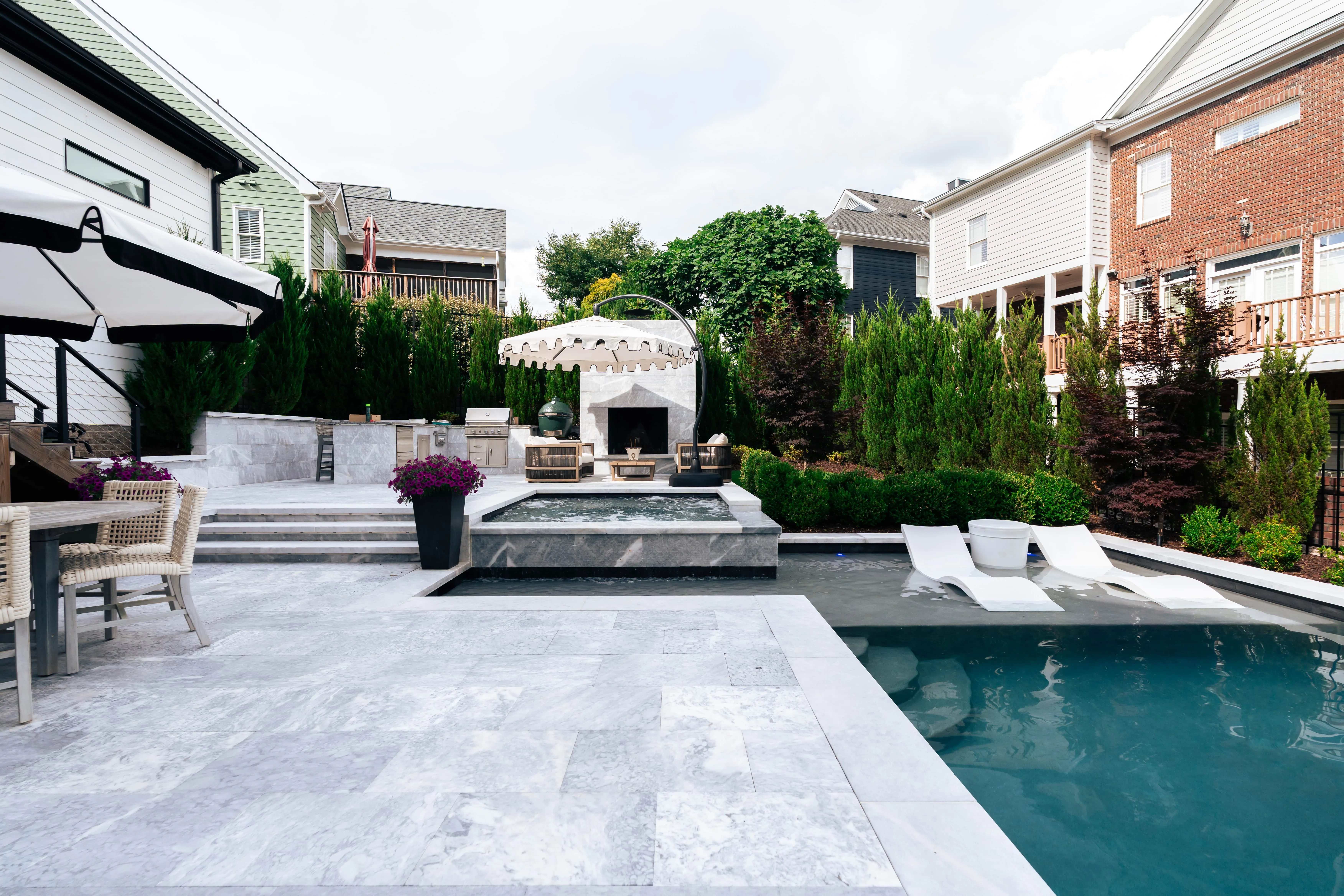 Wide-angle daytime view of a luxury rectangular inground swimming pool featuring an integrated concrete spa, a large light gray stone paver deck patio with modern lounge chairs, and a contemporary dark-accented pool house built by an outdoor living company in Raleigh, NC.