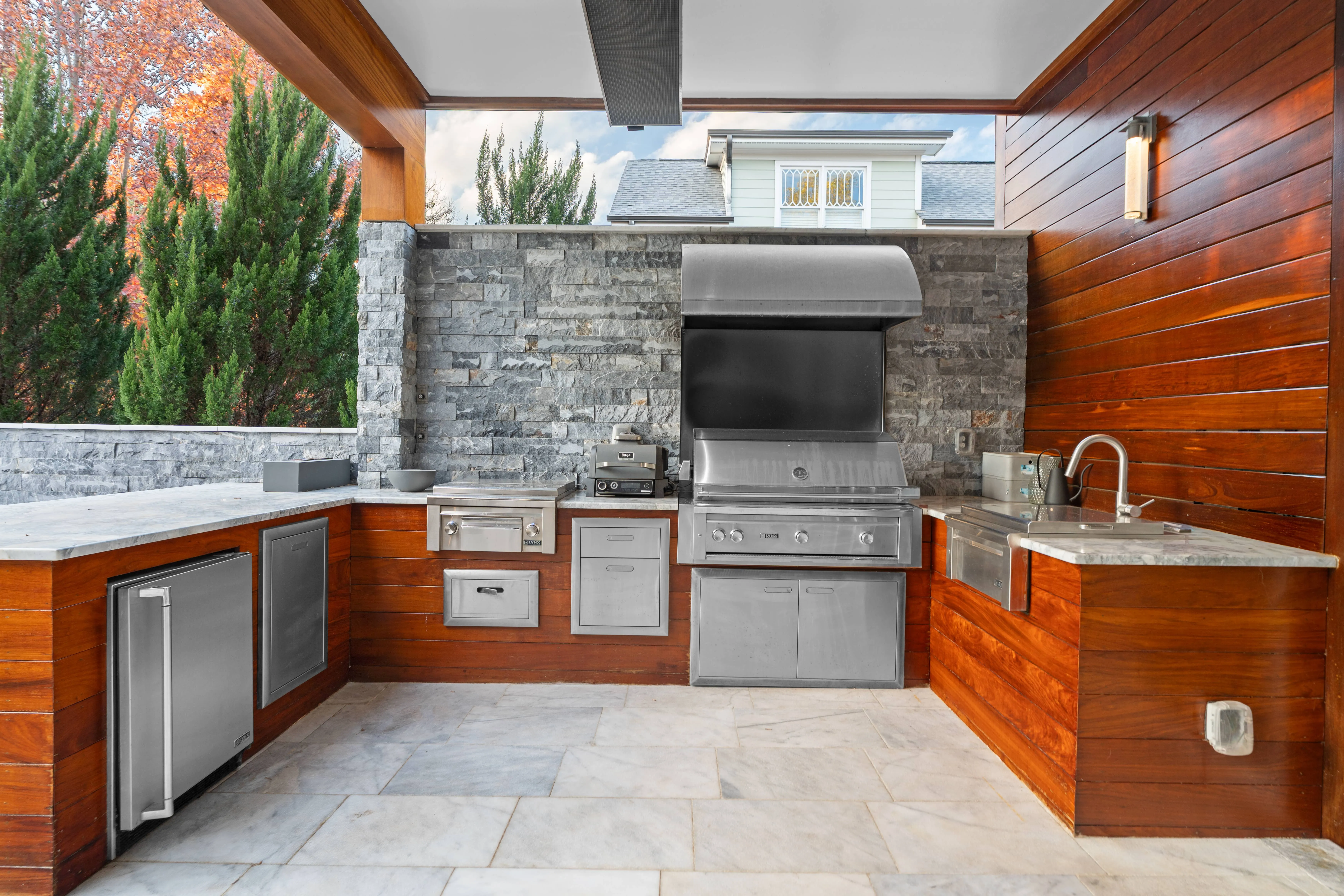 Luxury custom outdoor kitchen featuring a built-in stainless steel grill, under-counter refrigerator, and stone accent walls beneath a modern covered patio pavilion built next to an inground swimming pool by an outdoor living company in Raleigh, NC.