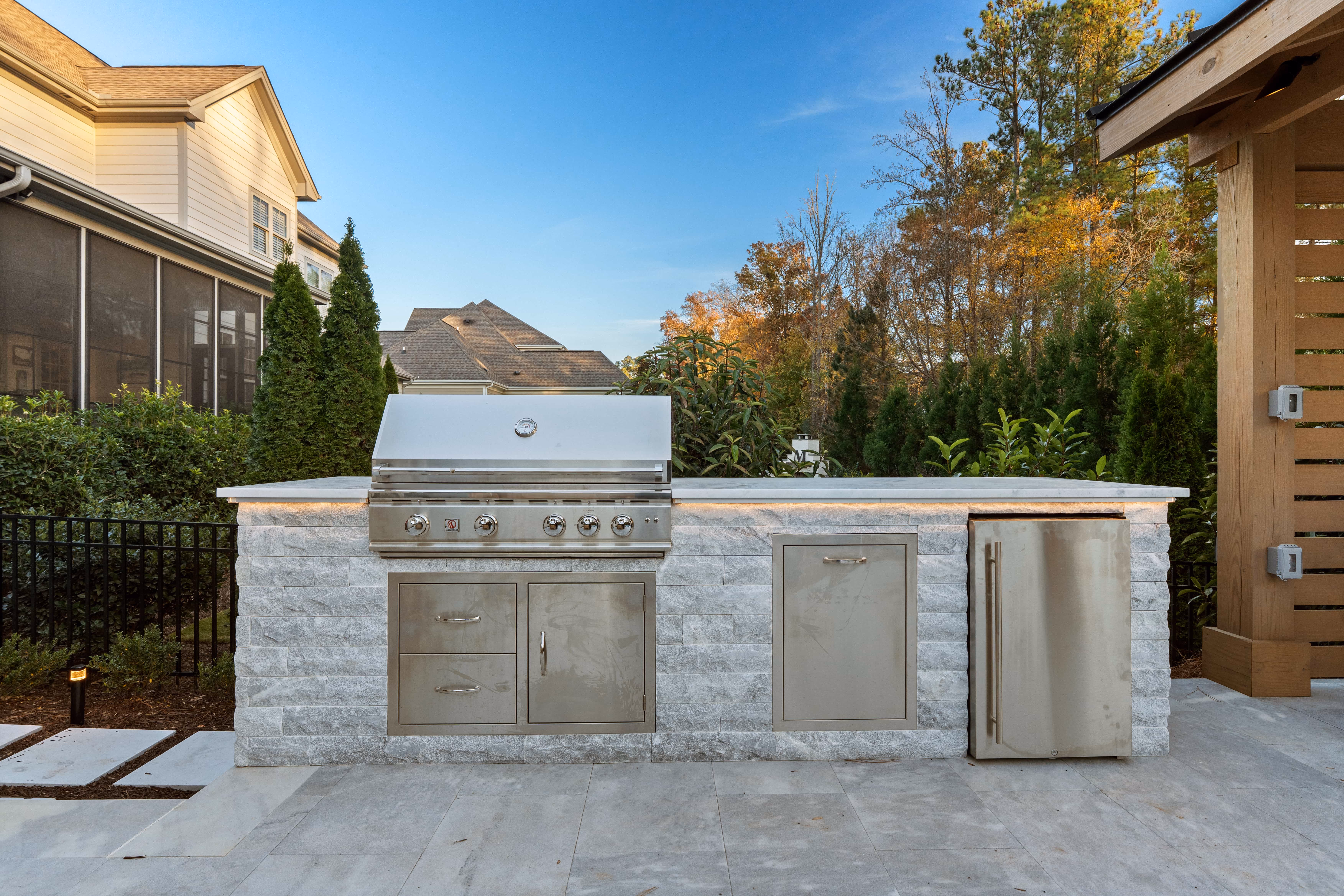 Straight-on daytime view of a luxury custom outdoor grill station featuring a built-in stainless steel gas grill, matching access drawers, and a compact refrigerator set into light stone veneer siding built by an outdoor living company in Raleigh, NC.