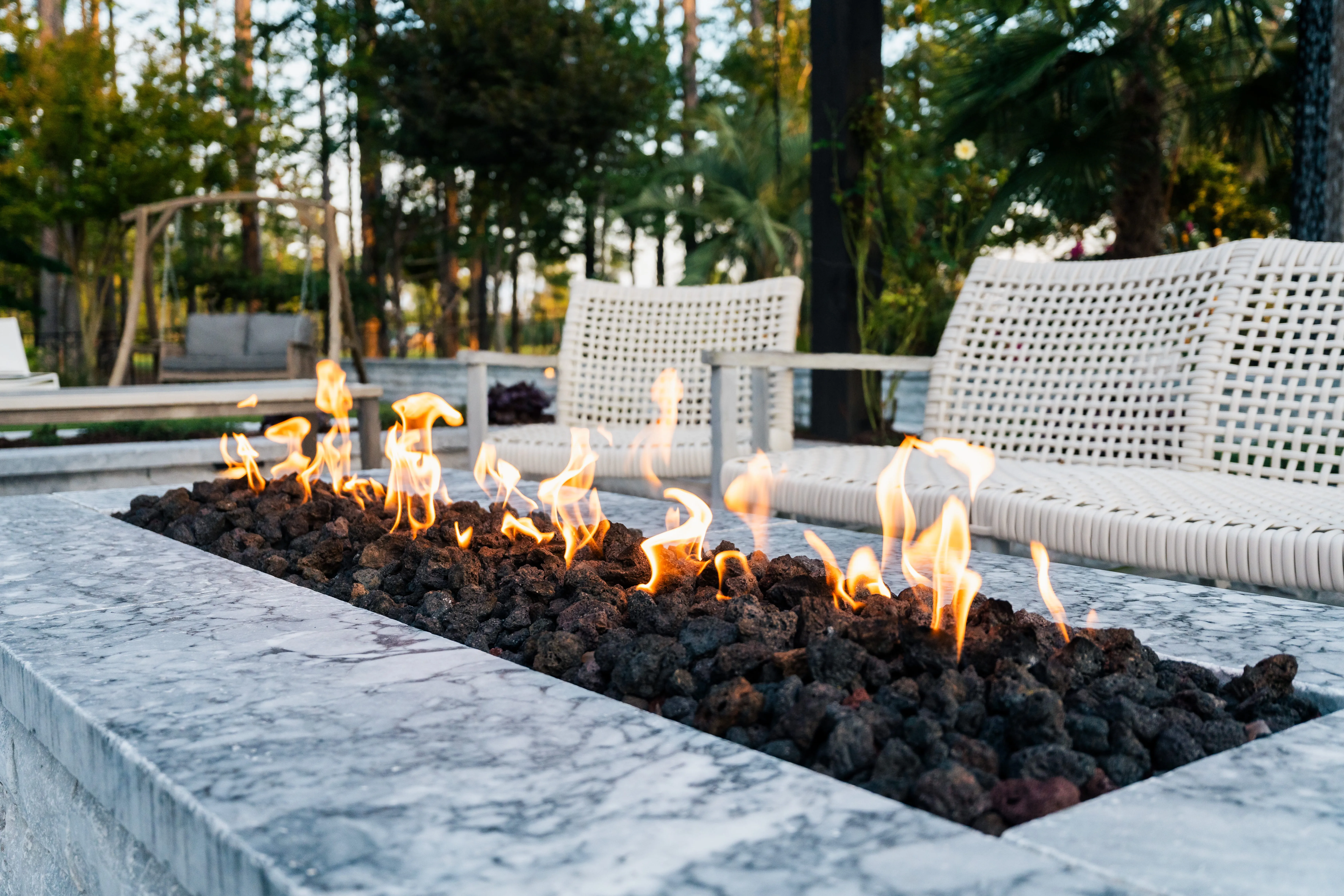 Close-up twilight view of a luxury modern outdoor linear fireplace with active flames, built alongside stylish white mesh patio lounge chairs by an outdoor living company in Raleigh, NC.