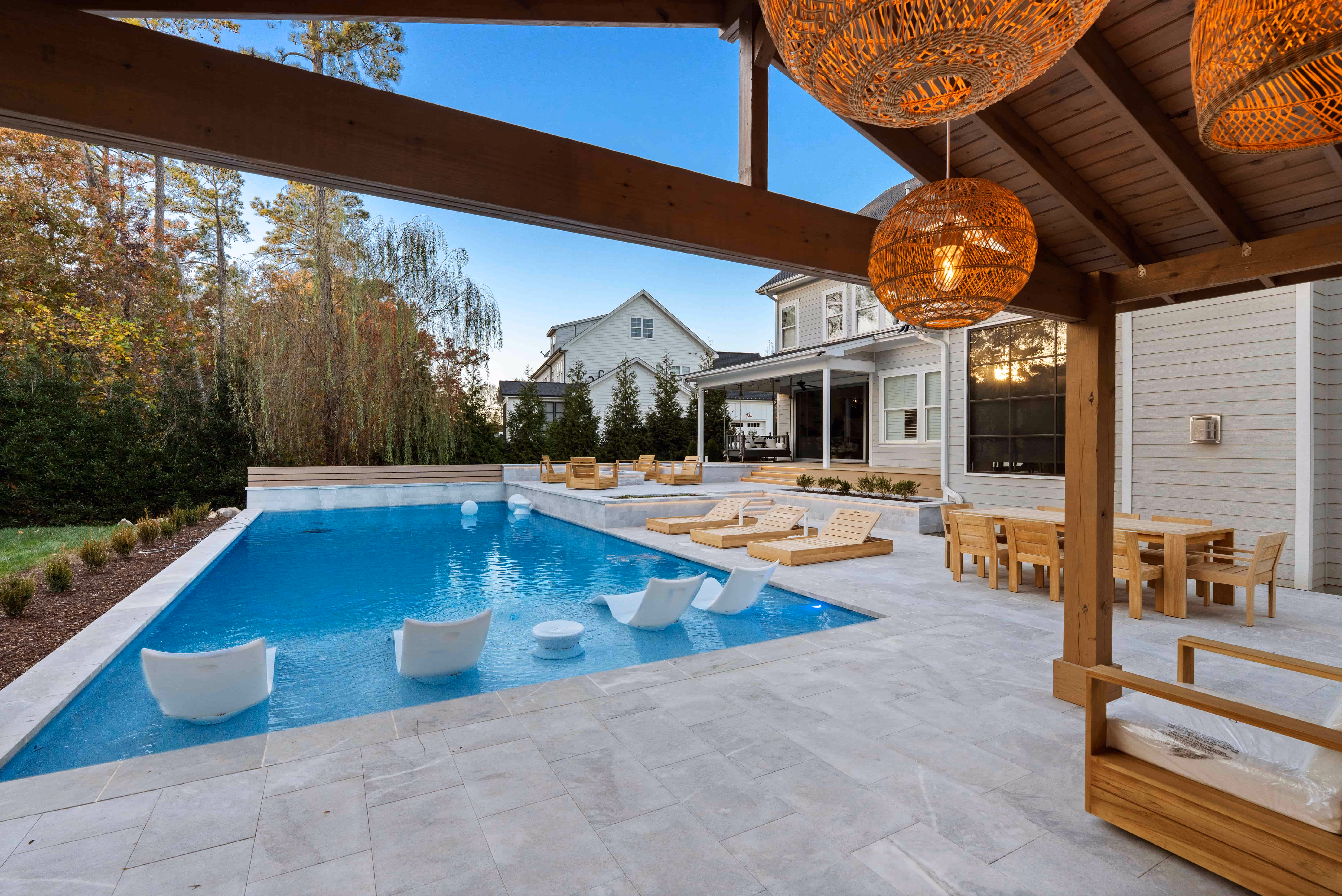 Wide-angle backyard view of a luxury rectangular swimming pool with white in-pool ledge loungers, a spacious stone paver patio deck, and a grand wood pavilion cabana featuring hanging basket lights built by an outdoor living company in Raleigh, NC.