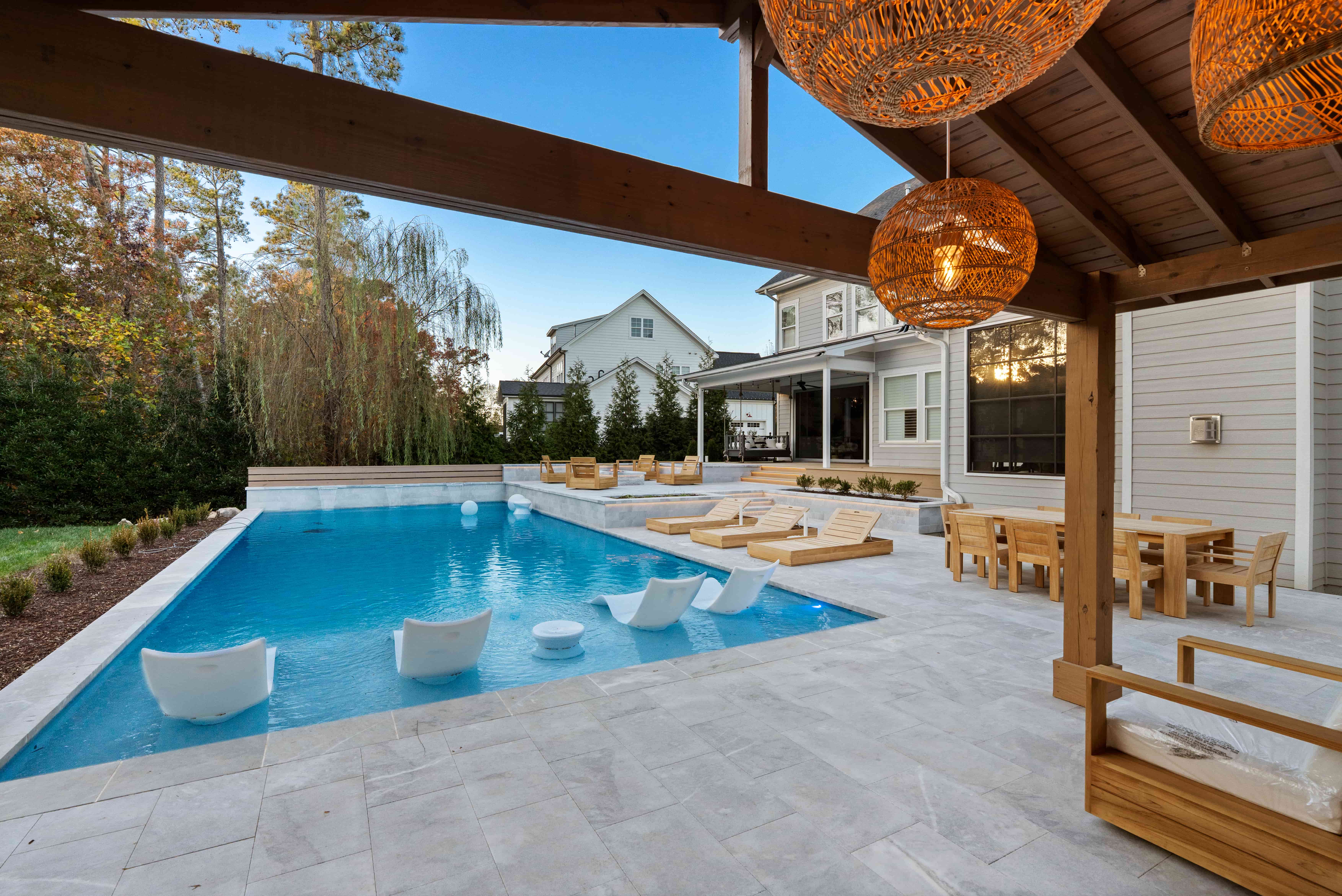 Wide-angle backyard view of a luxury rectangular swimming pool with white in-pool ledge loungers, a spacious stone paver patio deck, and a grand wood pavilion cabana featuring hanging basket lights built by an outdoor living company in Raleigh, NC.