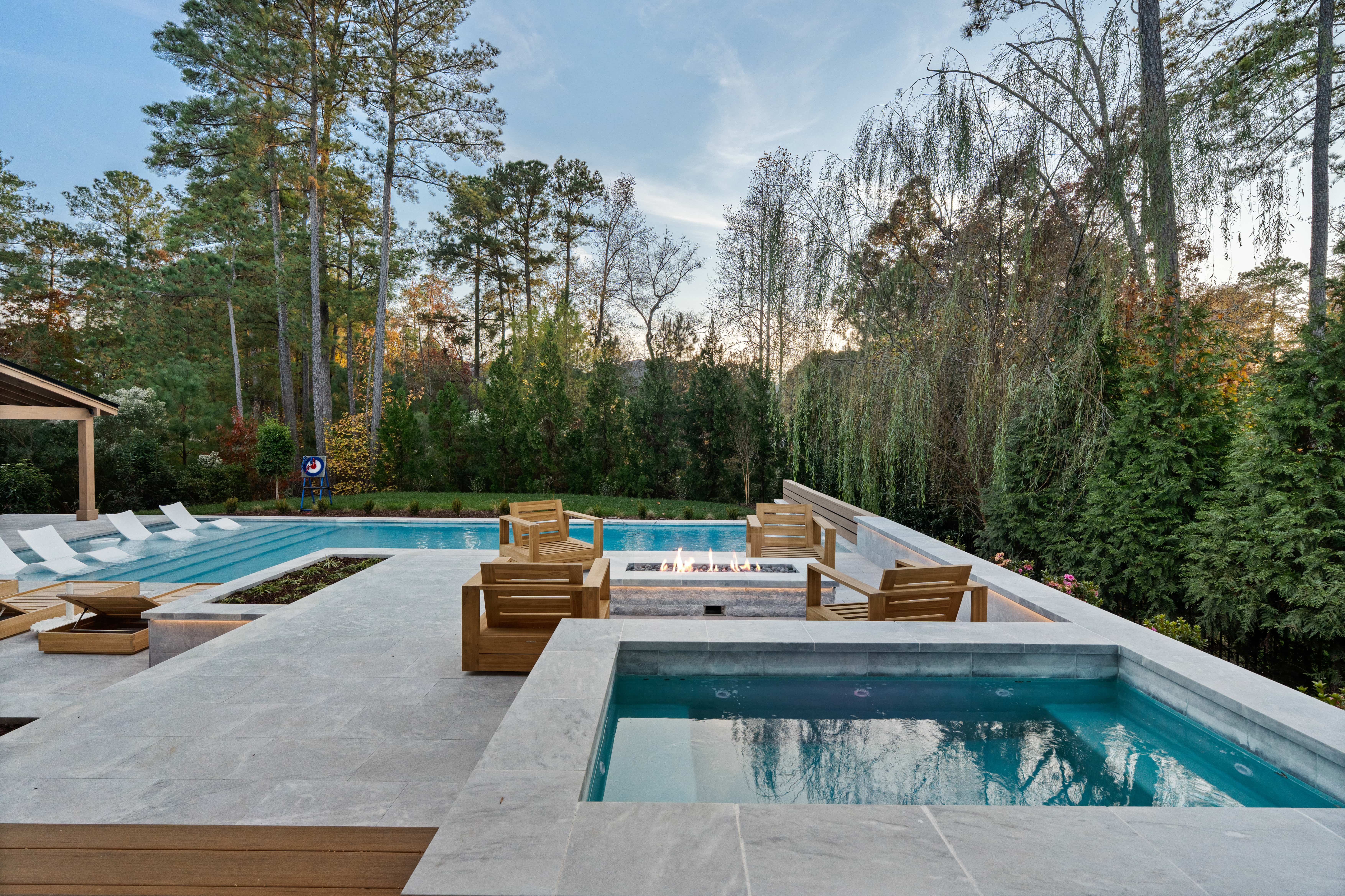 High-angle view of a luxury residential backyard transformation featuring a custom rectangular swimming pool with an attached spa, a light stone paver patio decking layout, a dark-roofed pool house cabana, and a dense backdrop of mature pine trees built by an outdoor living company in Raleigh, NC.