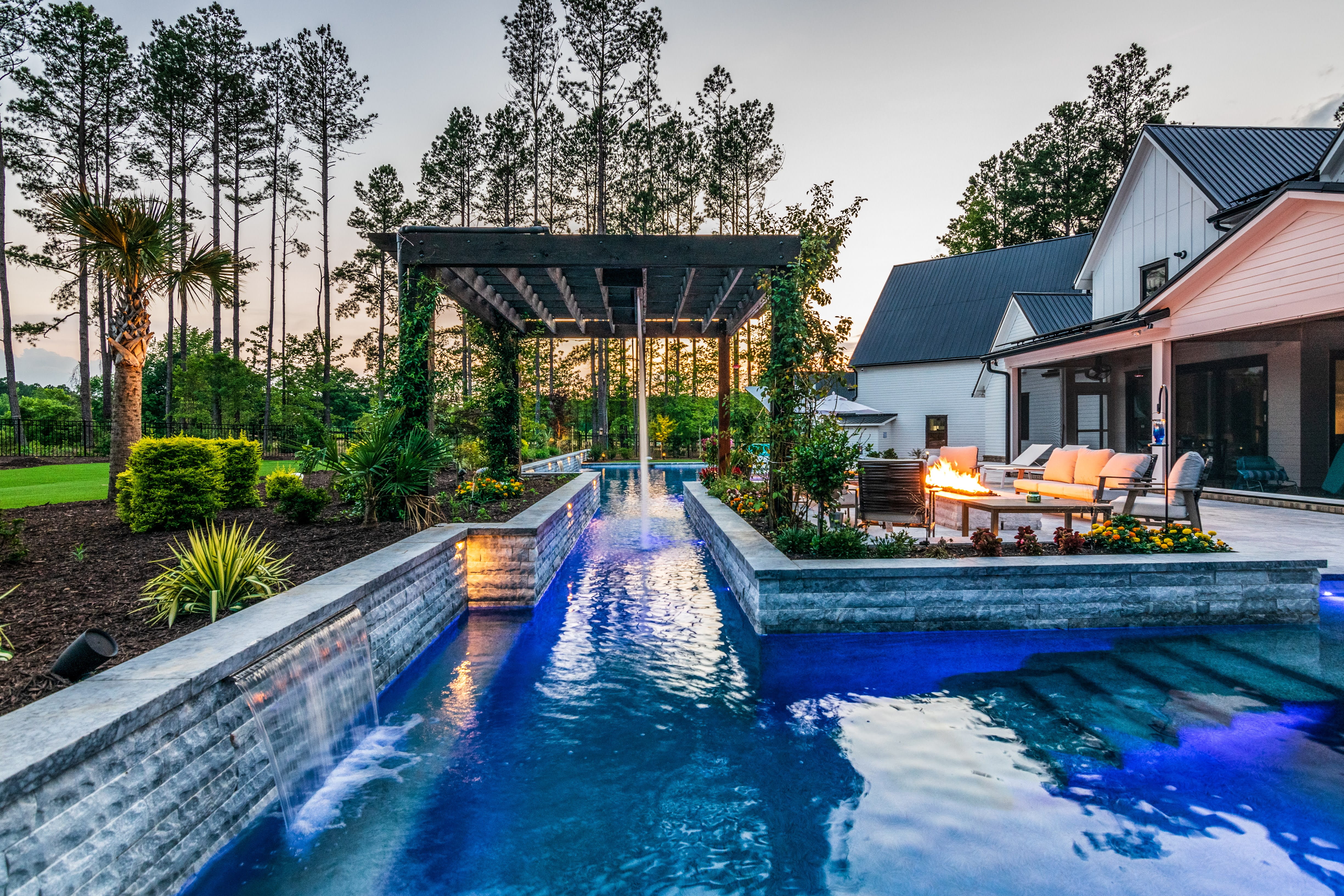 High-angle aerial view of a custom rectangular swimming pool with light stone paver decking, luxury lounge chairs, a modern dark-roofed pool house, and mature trees built by an outdoor living company in Raleigh, NC.