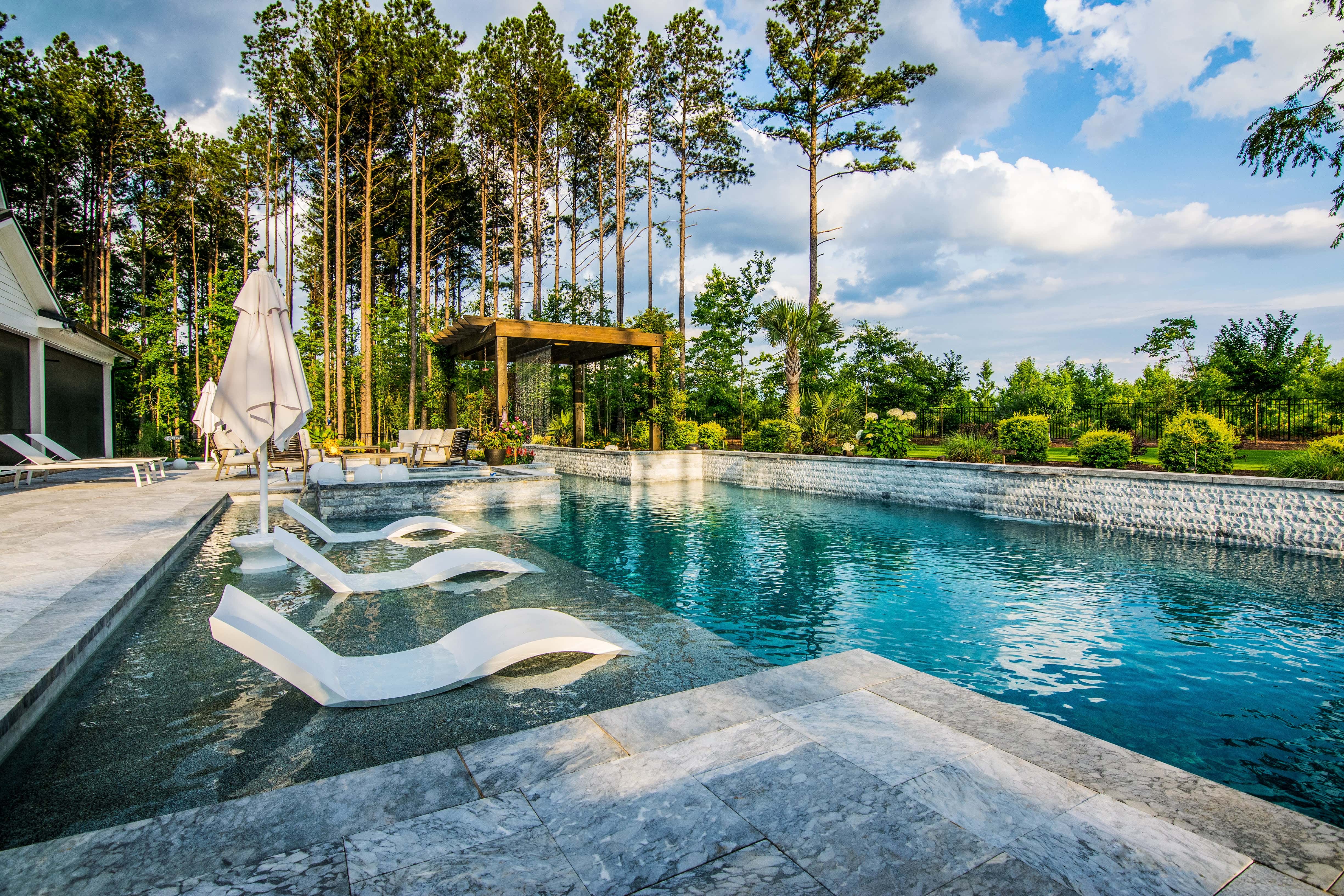 Wide-angle view of a luxury rectangular swimming pool featuring white in-pool ledge lounger chairs, light stone paver decking, an elegant wood pavilion pergola, and a dense backdrop of mature pine trees built by an outdoor living company in Raleigh, NC.