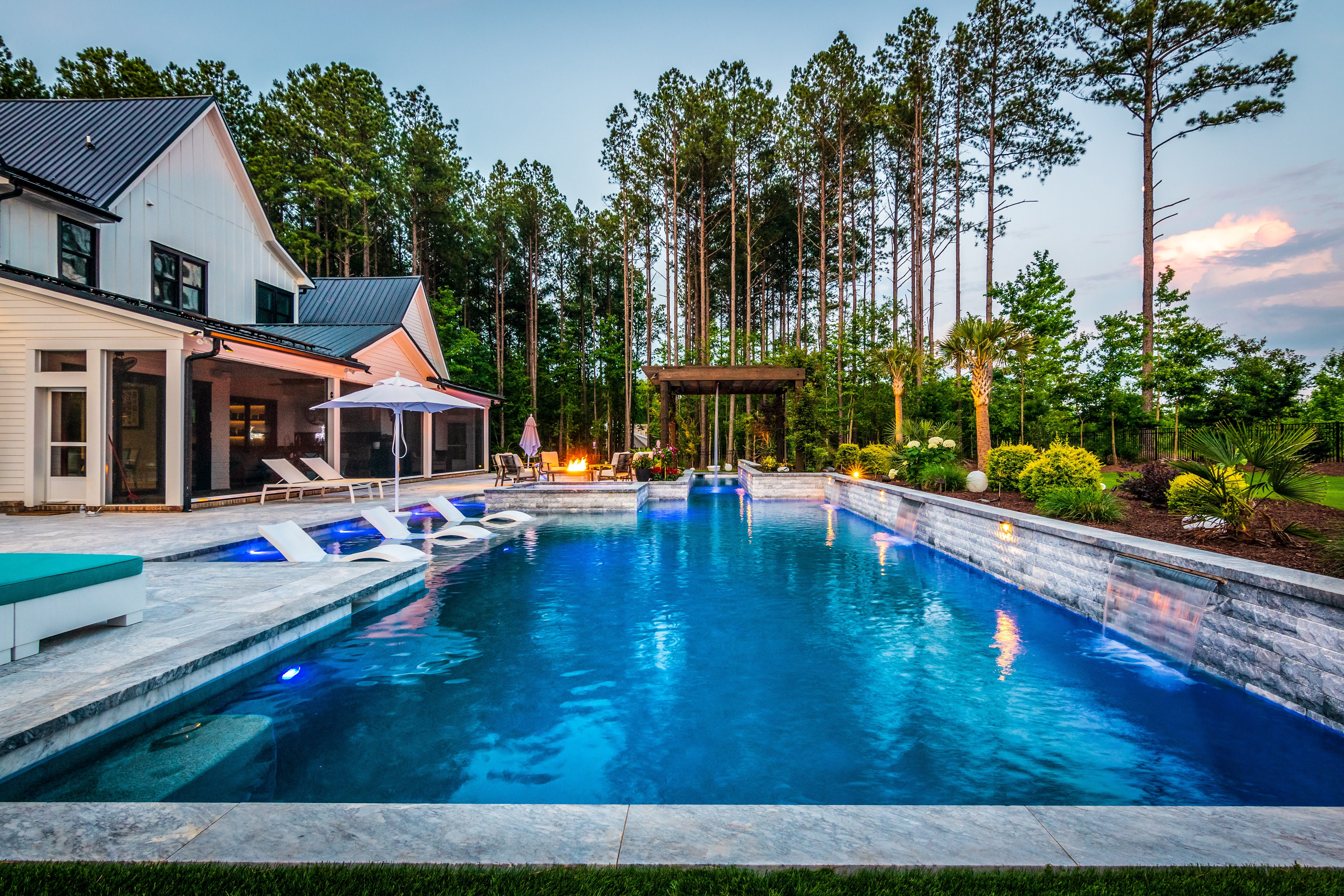 High-angle view of a luxury residential backyard featuring a rectangular swimming pool, custom light gray stone paver pool decking, a modern black pergola lounging area, and a lush background of mature trees built by an outdoor living company in Raleigh, NC.