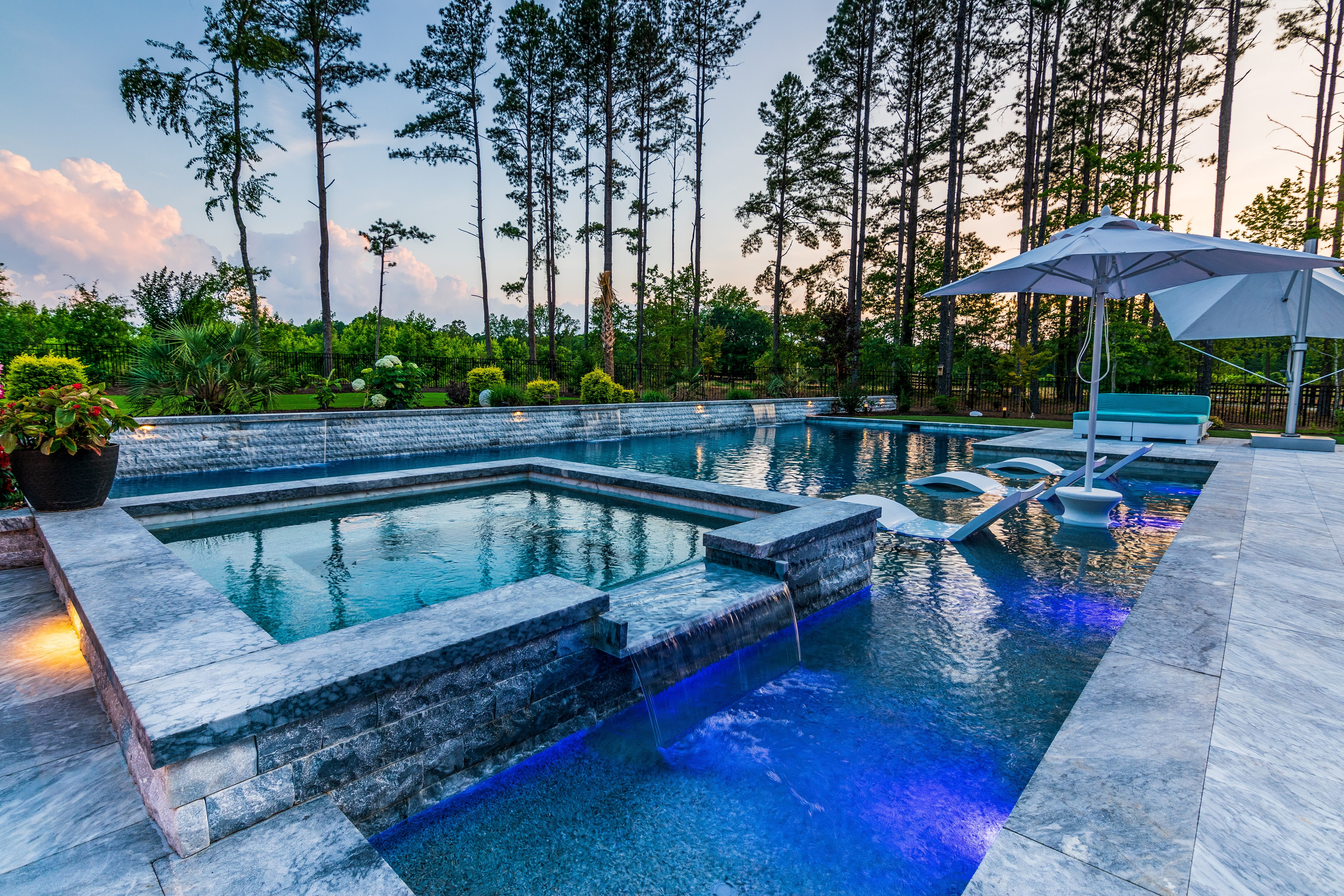 Wide-angle panoramic view of a luxury residential backyard featuring a custom swimming pool, a light stone paver patio with a white umbrella lounge area, and a lush backdrop of mature pine trees designed by an outdoor living company in Raleigh, NC.