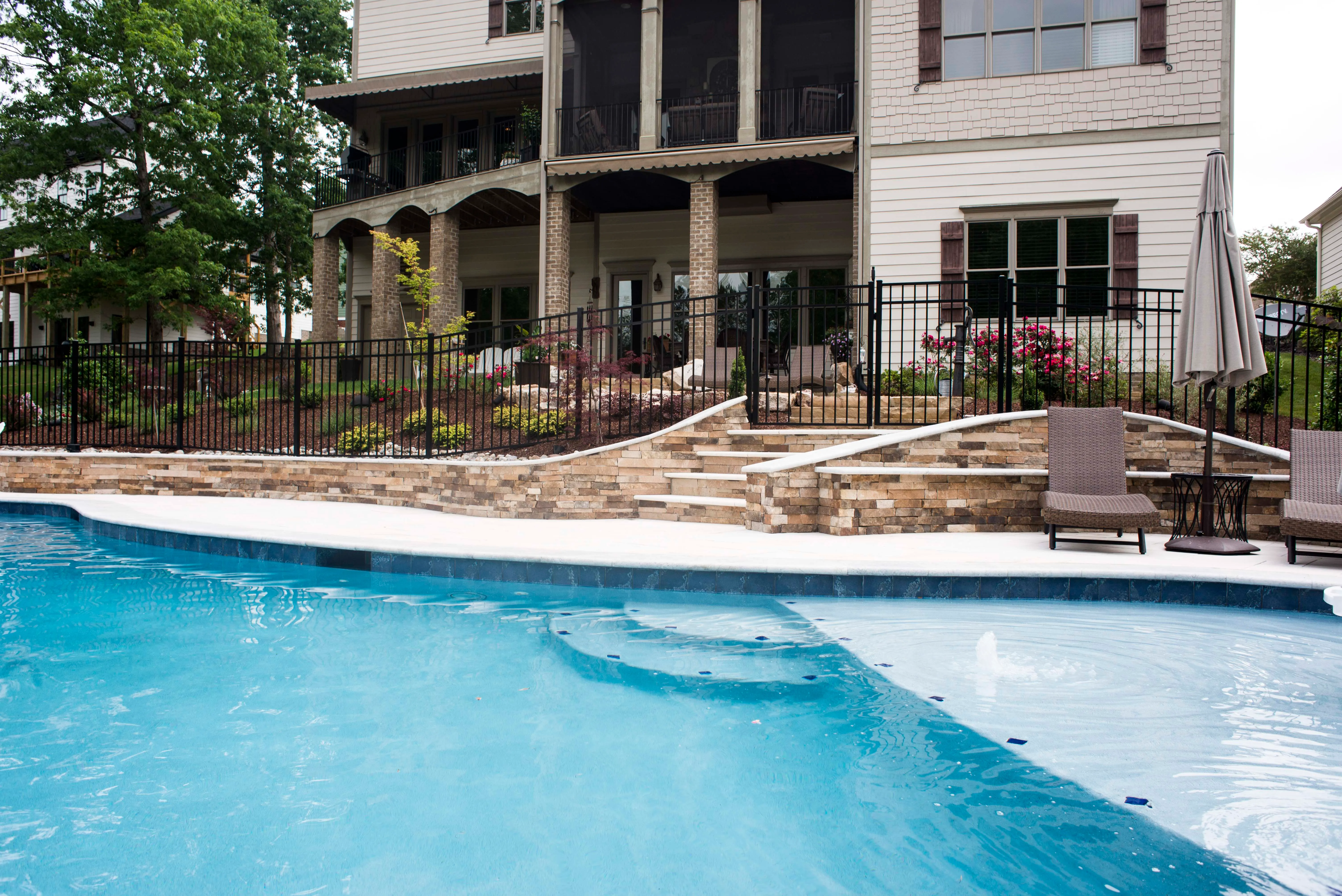 Luxury custom swimming pool design featuring clear blue water, a stone patio surround, and a wrought iron safety fence with an elegant two-story brick home backdrop built by a custom pool builder in Raleigh, NC.