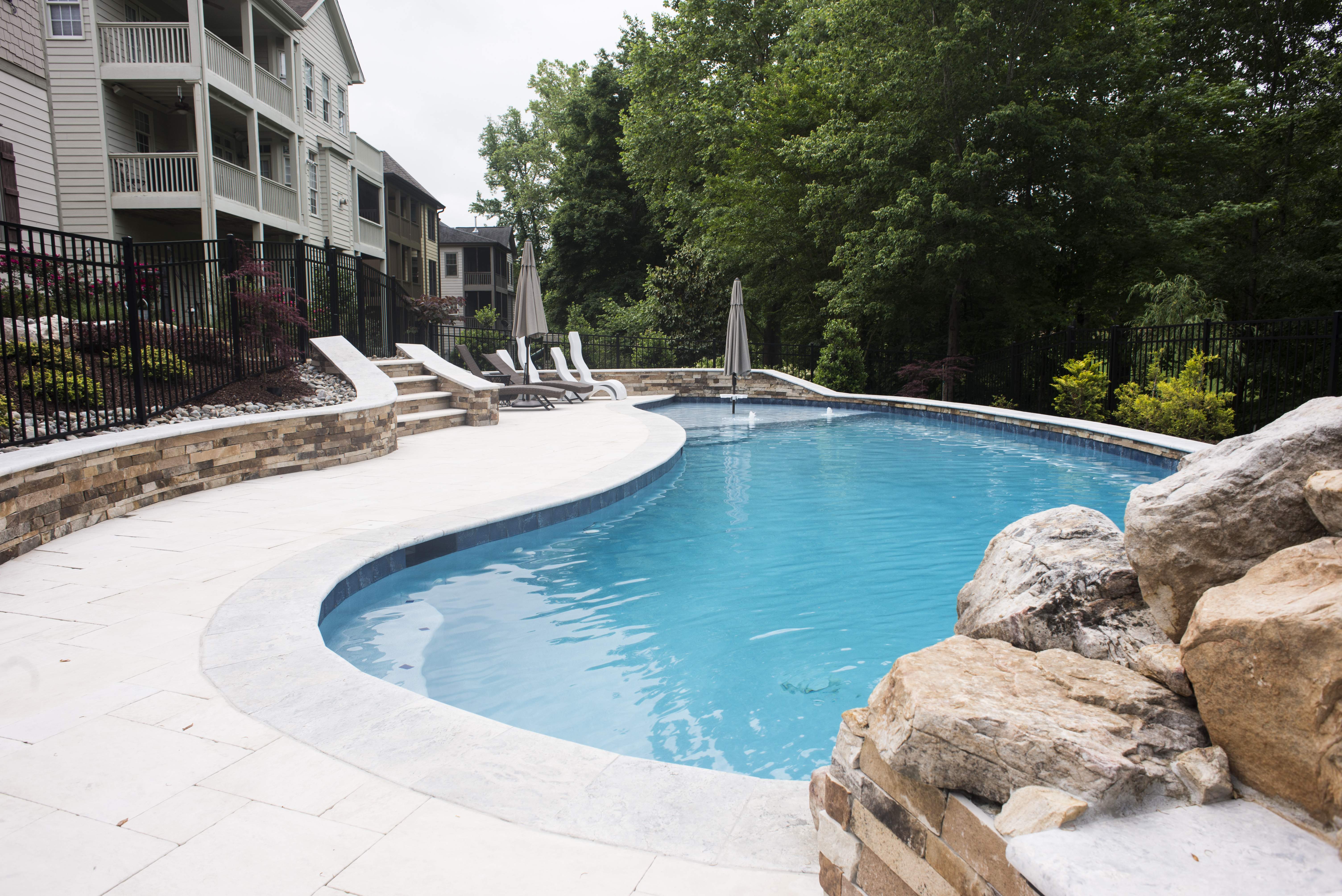 Custom lagoon swimming pool design featuring a natural stone rock waterfall, a smooth concrete pool deck, and lush backyard landscaping built by a pool builder in Raleigh, NC.