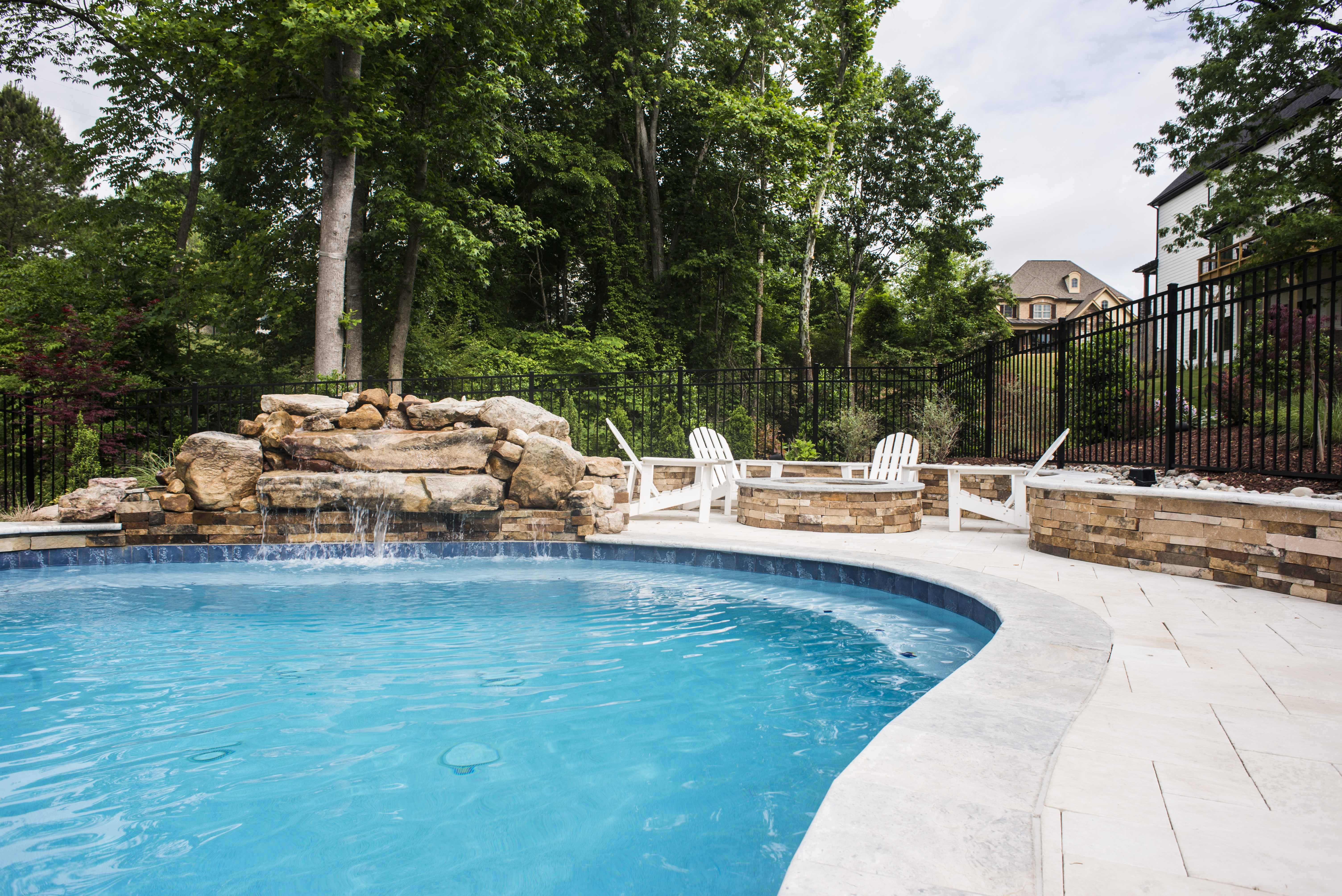 Custom-built swimming pool featuring a natural stone rock waterfall, an adjacent stone fire pit patio with white lounge chairs, and a lush tree canopy backdrop built by a custom pool builder in Raleigh, NC.