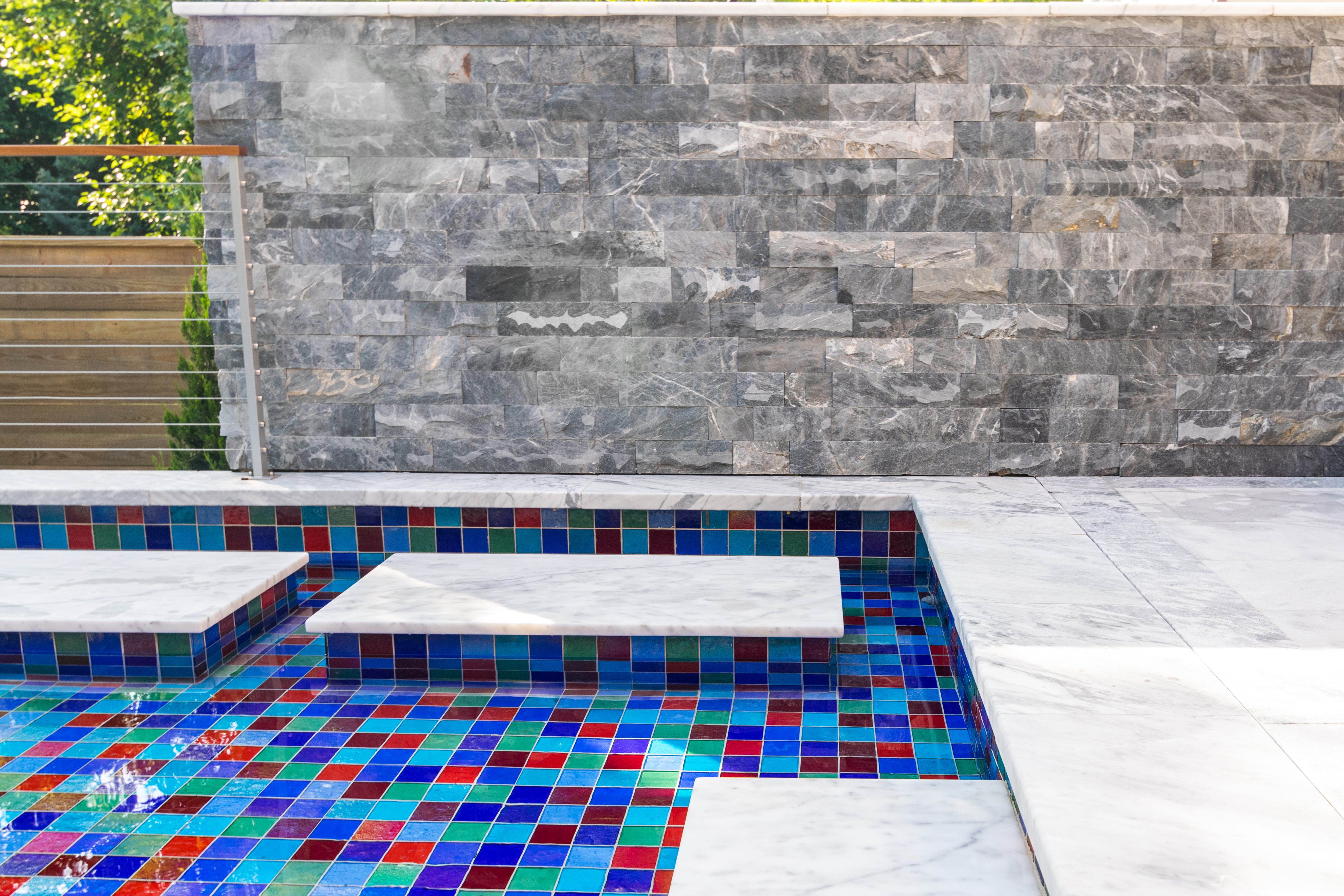 Modern geometric swimming pool featuring colorful blue and red mosaic glass tiling and a contemporary stone accent wall built by a custom pool builder in Raleigh, NC.