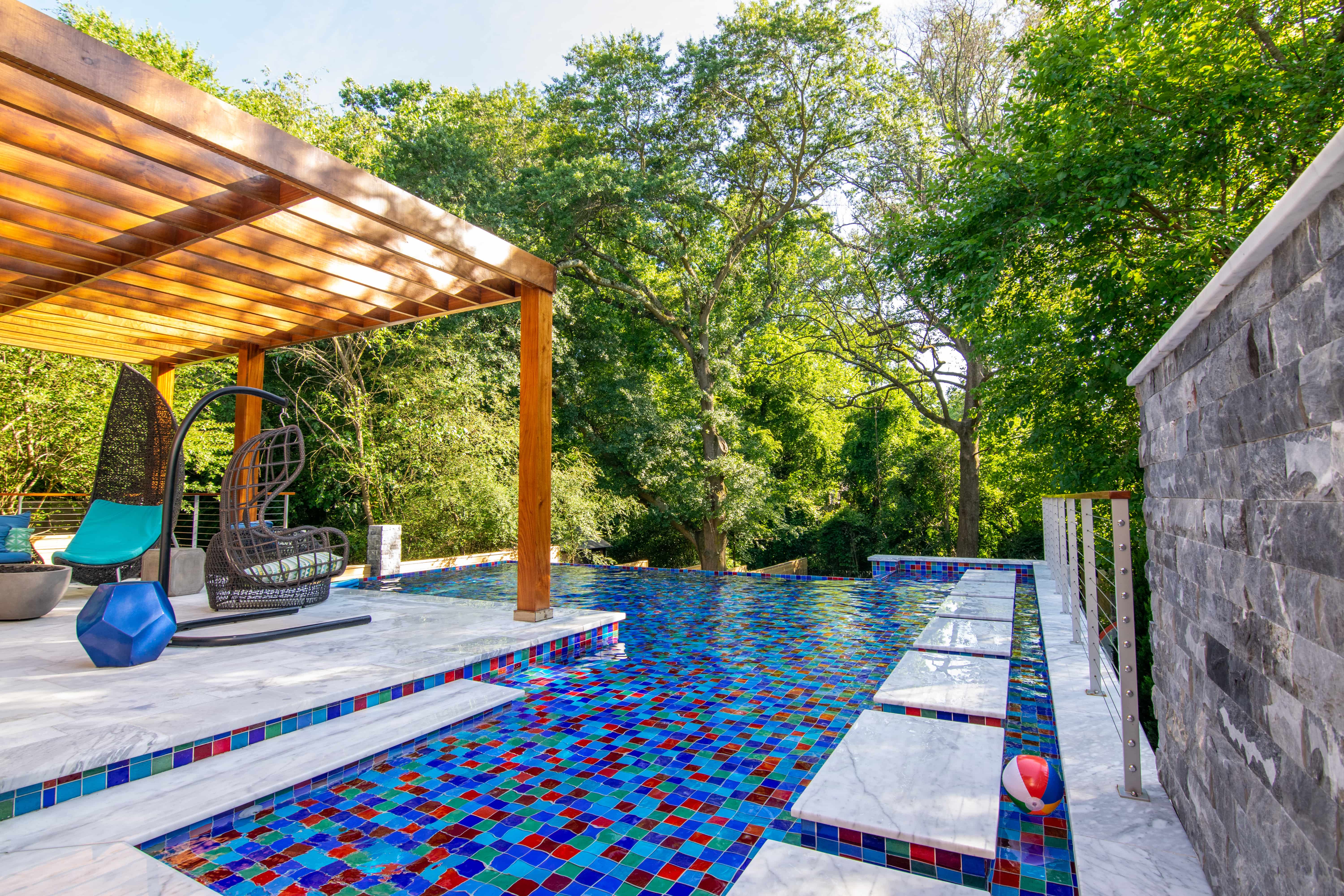 Luxury geometric swimming pool with vibrant mosaic glass tiling and an adjacent wooden patio pergola built by a custom pool builder in Raleigh, NC.