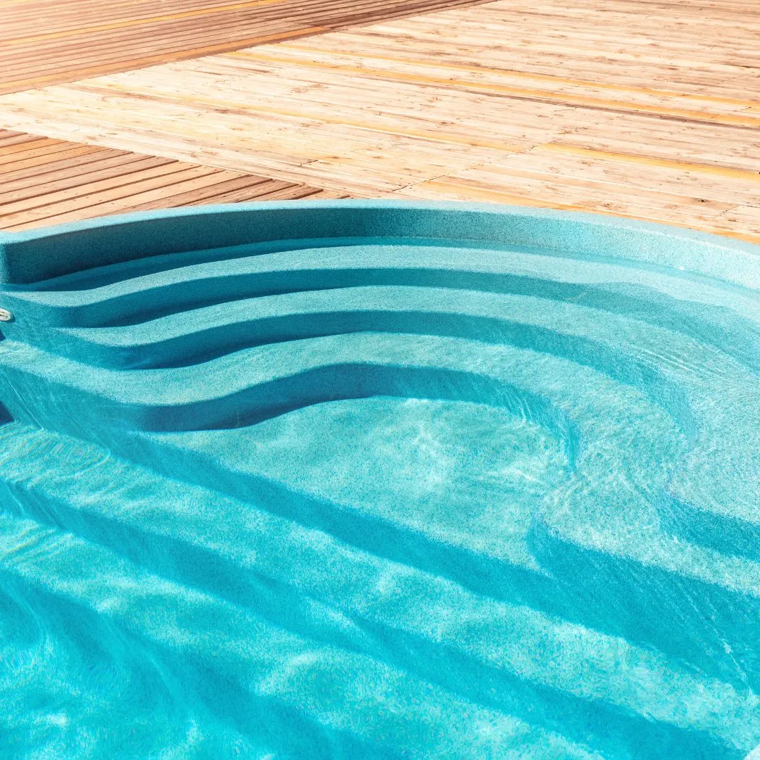 Custom inground swimming pool entry featuring curved steps, shallow sun shelf seating area, clear turquoise water, and wood deck surround in Fuquay-Varina, NC.
