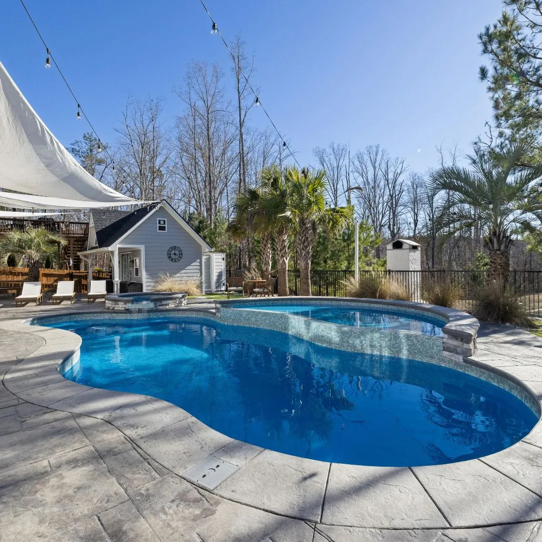 Custom freeform inground swimming pool featuring deep blue water, light stone patio surround, and tropical palm tree landscaping in Fuquay-Varina, NC.