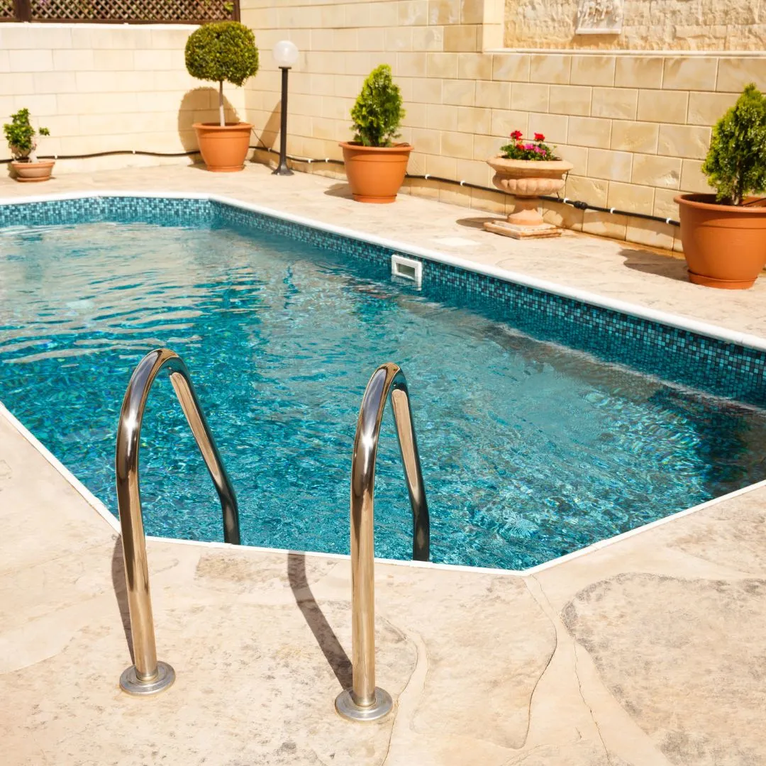 Custom inground swimming pool featuring stainless steel entry ladder handrails, tan patio tile surround, and potted plants along a stone retaining wall in Garner, NC.