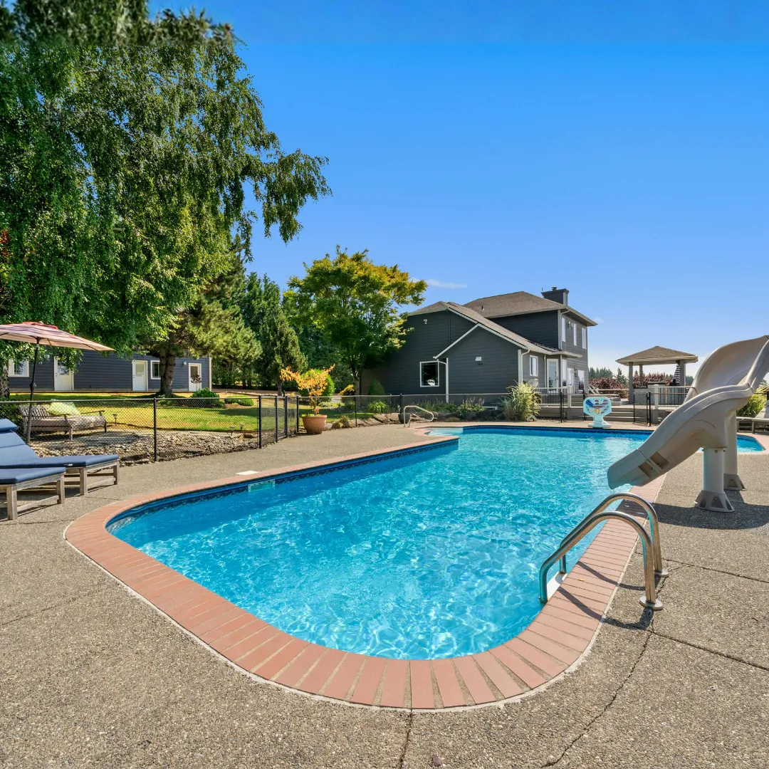 Custom curved inground swimming pool featuring a backyard water slide, brick patio coping, and surrounding lawn landscaping in Raleigh, NC.