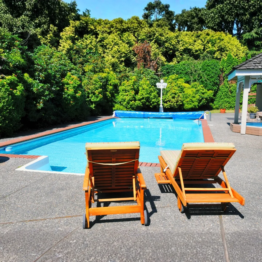 Custom inground swimming pool featuring wooden lounge chairs on a concrete patio surrounded by lush backyard landscaping in Raleigh, NC.
