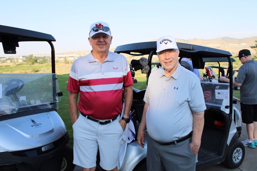 Two men standing in front of golf carts on a sunny golf course with hills in the background.