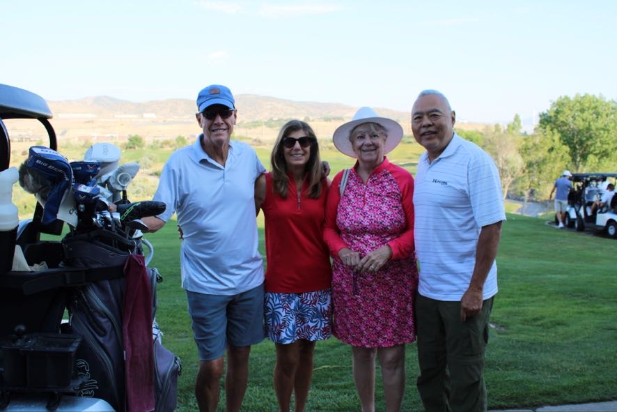 Four adults posing together outdoors on a golf course with golf carts and clubs nearby.