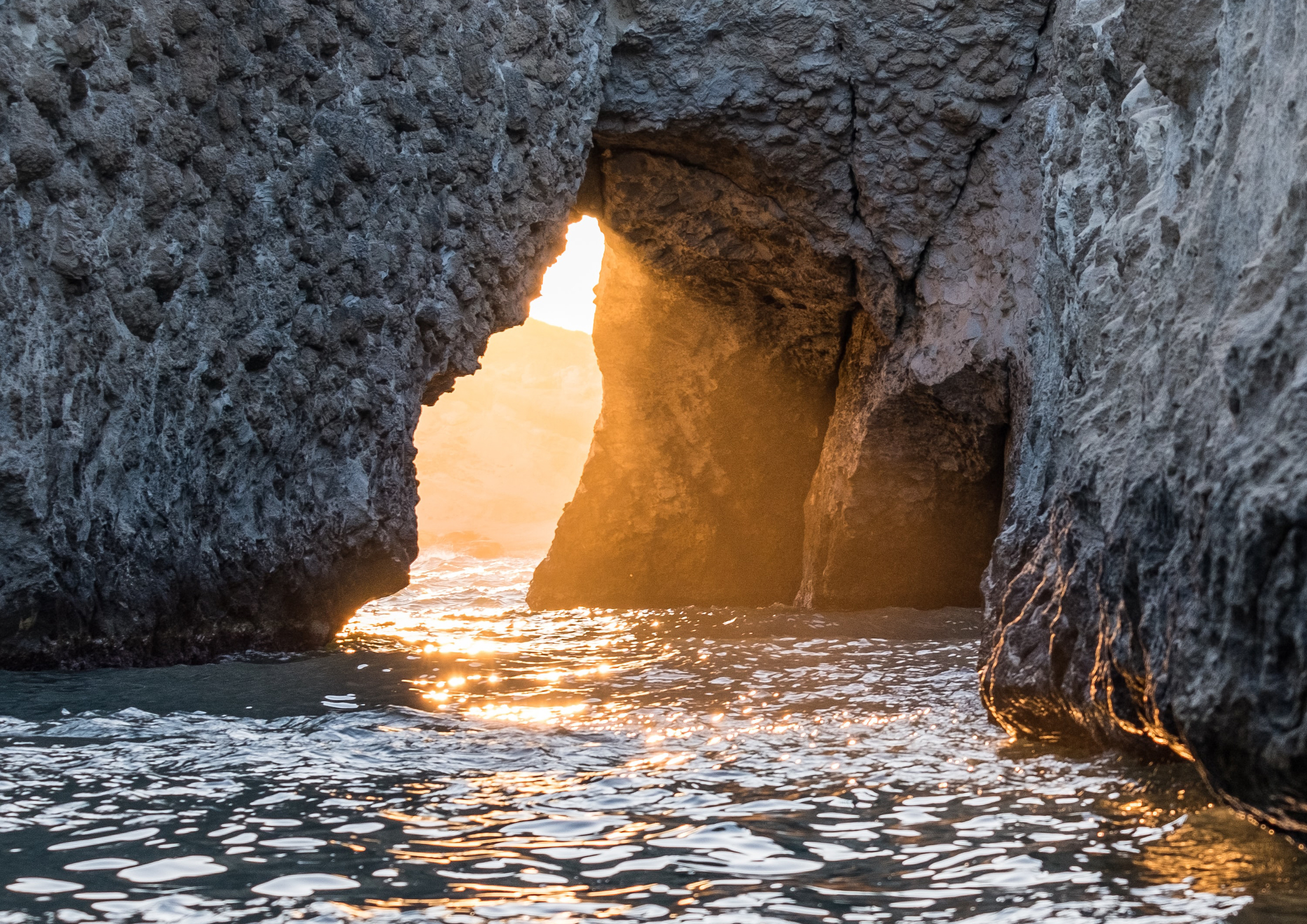 Warm light pours through an opening in the rocks, reflecting off the soft ocean waves.