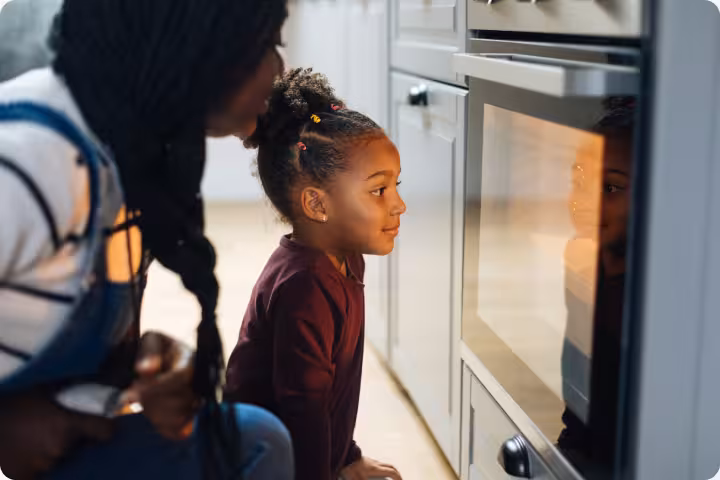 Young girl and woman watching food inside the oven in a kitchen.