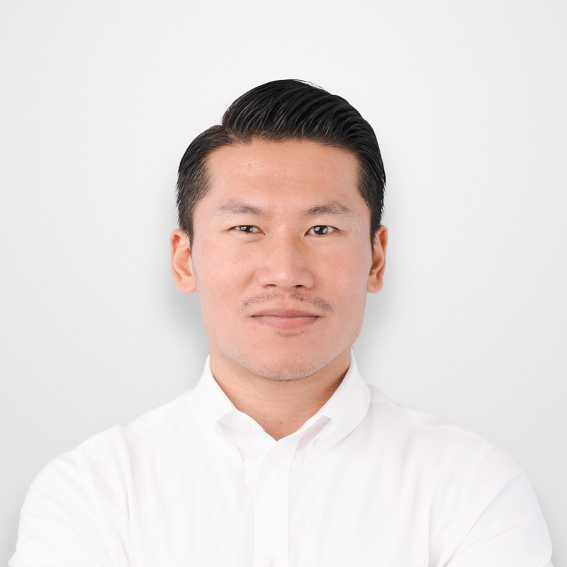 白いシャツを着た黒髪の男性が微笑んで正面を見ている。