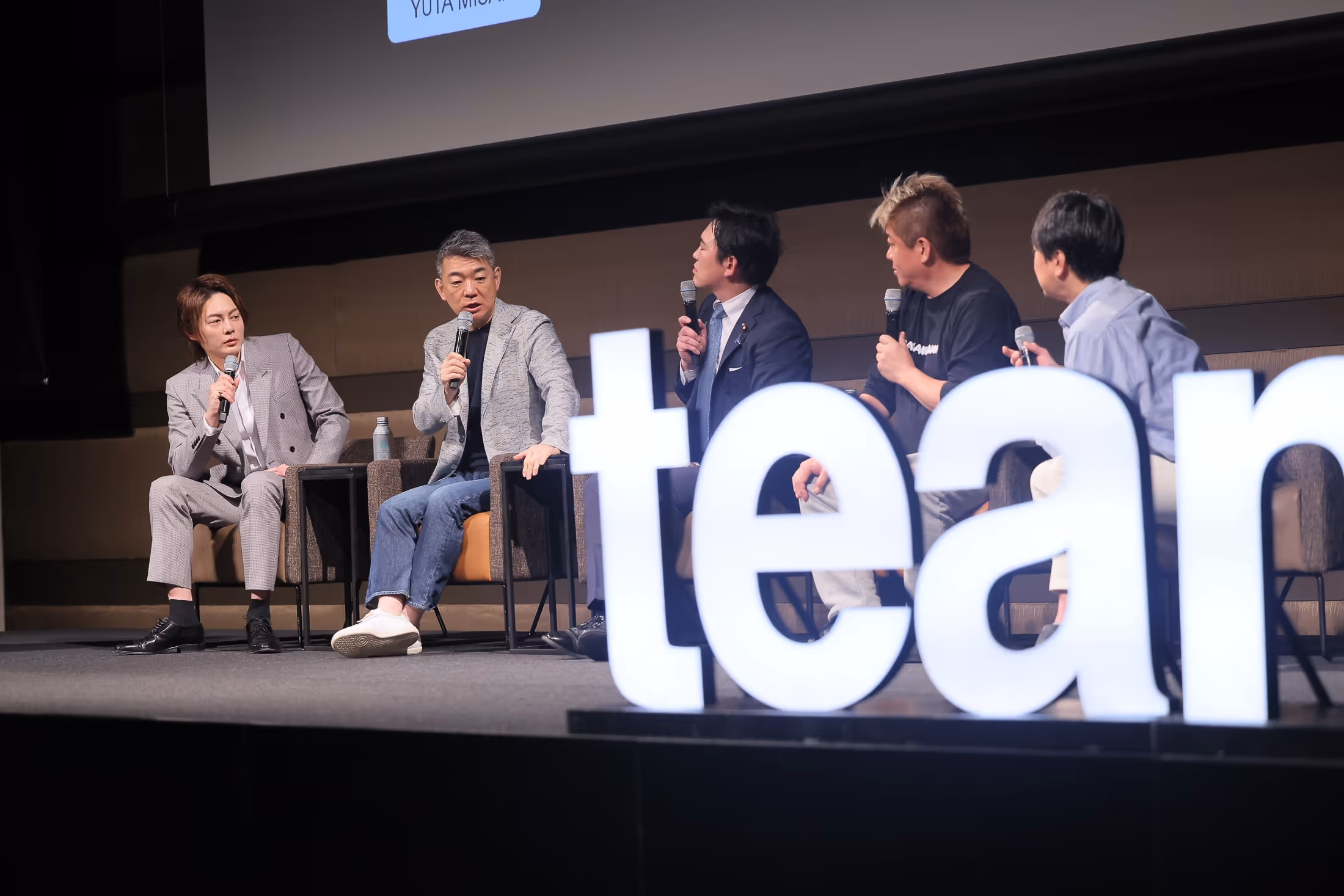 壇上でマイクを持って話す五人の男性が椅子に座り、前には大きな発光する『tea』の文字がある。