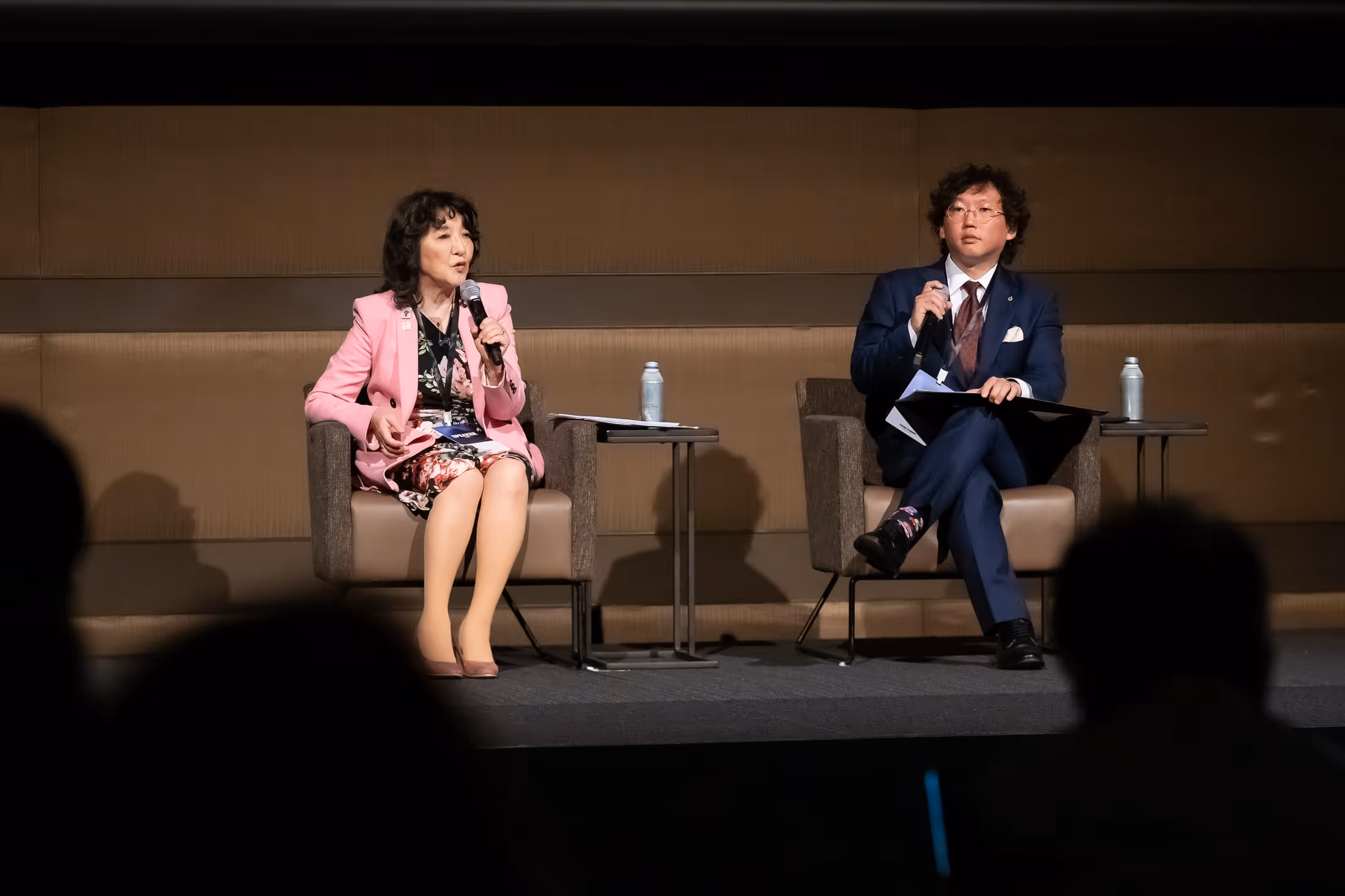 マイクを持ち討論しているピンクのジャケットを着た女性と青いスーツを着た男性が椅子に座っている。