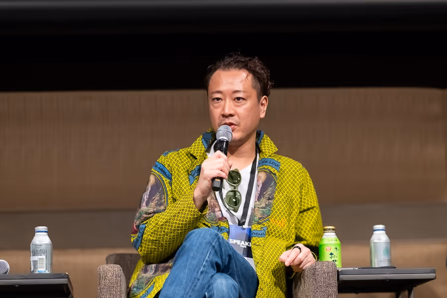 マイクを持ち話しているカジュアルな黄色いジャケットを着た男性。