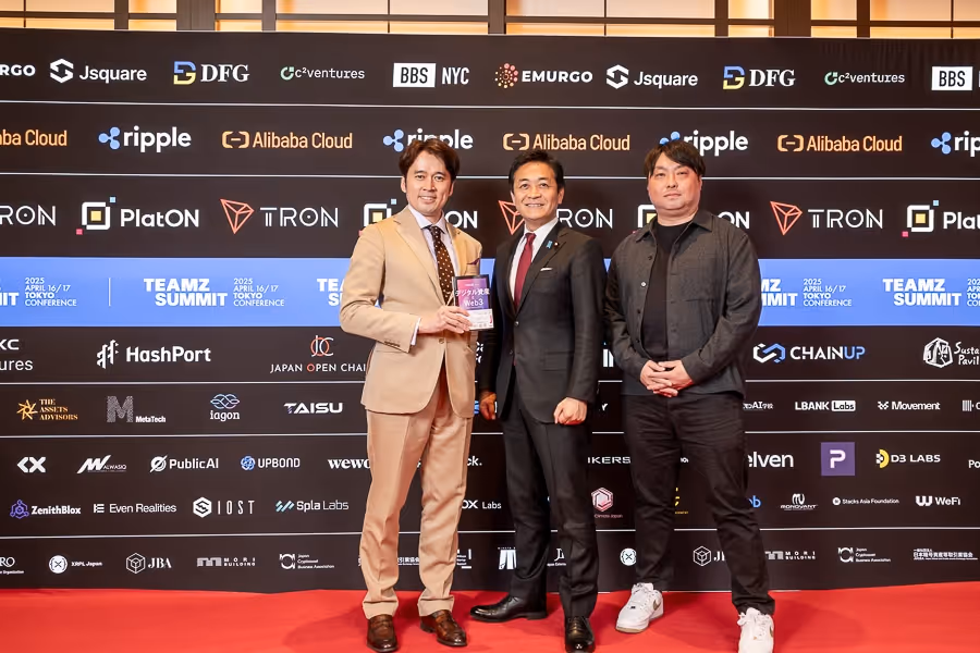 ビジネススーツを着た三人の男性がレッドカーペットの前でポーズを取り、中央の男性が賞を手にしている。