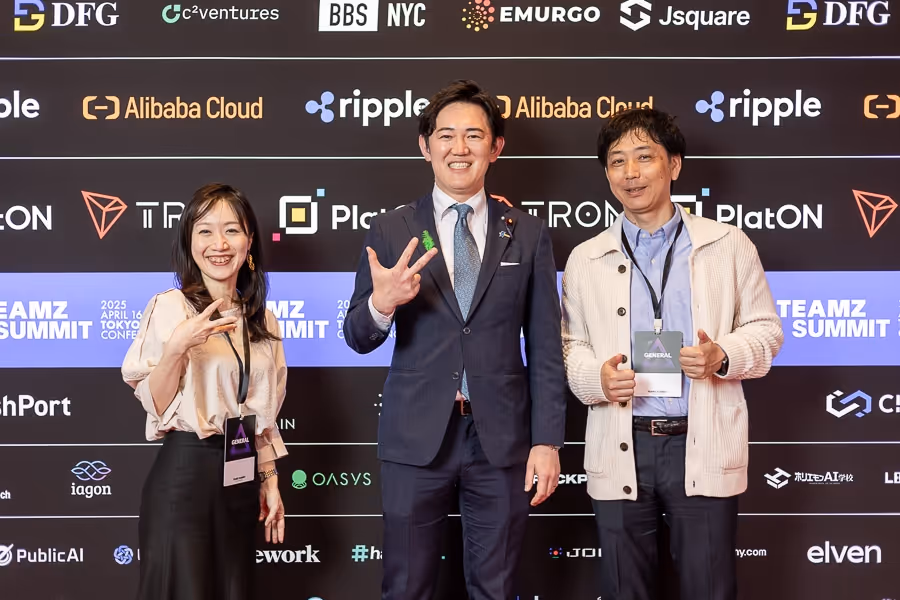 ビジネスカジュアルな服装の3人の男女が、イベントの背景を背に笑顔で手を挙げてポーズをとっている。