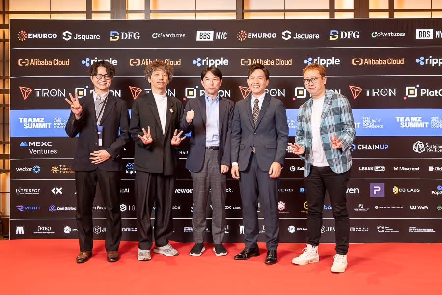 5人の男性が赤いカーペットに立ち、ビジネスカジュアルな服装でポーズを取っている。背景には複数の企業ロゴが並ぶ。