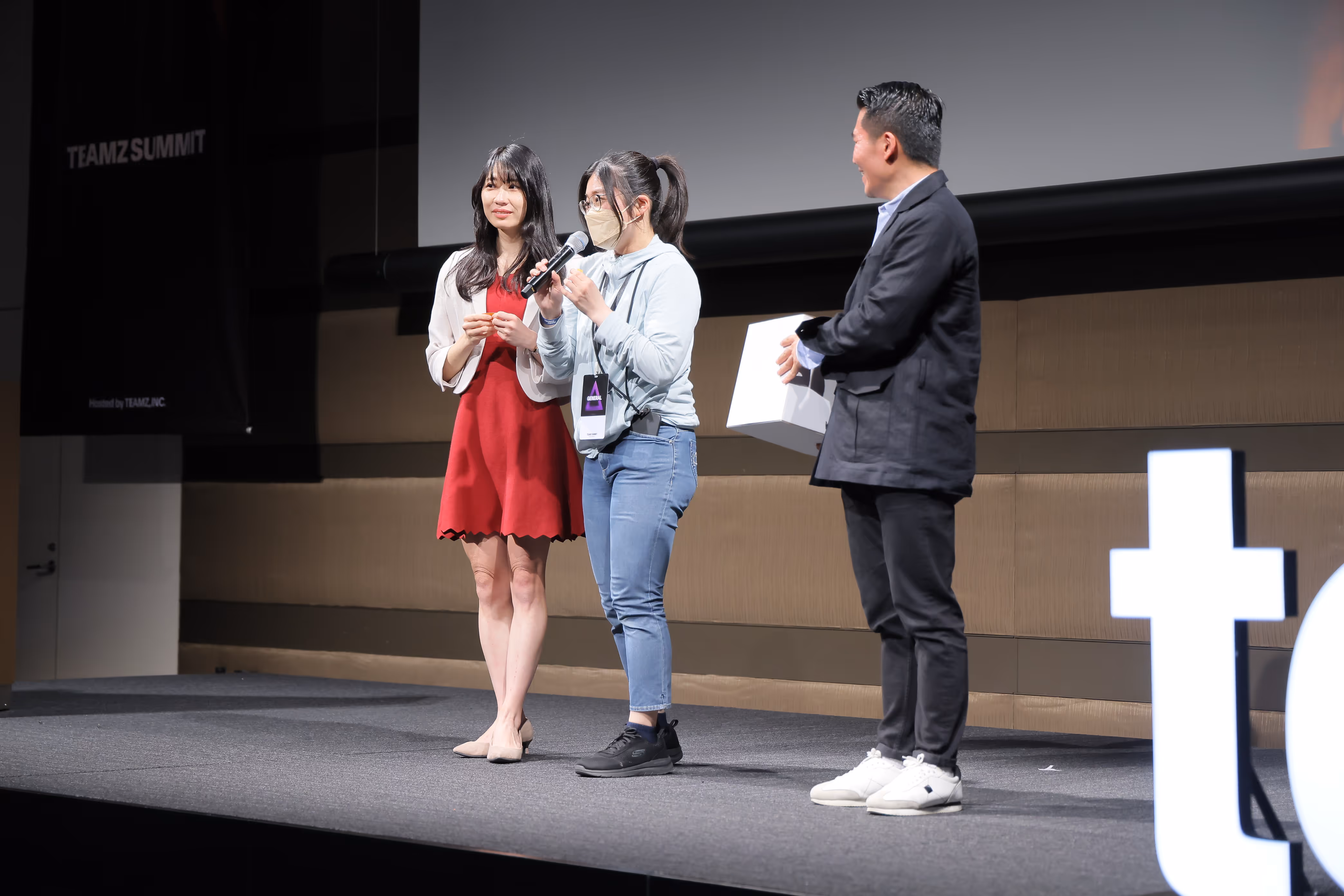 マイクを持ち話す女性と、赤いワンピースの女性、箱を持つ男性がステージに立っている。