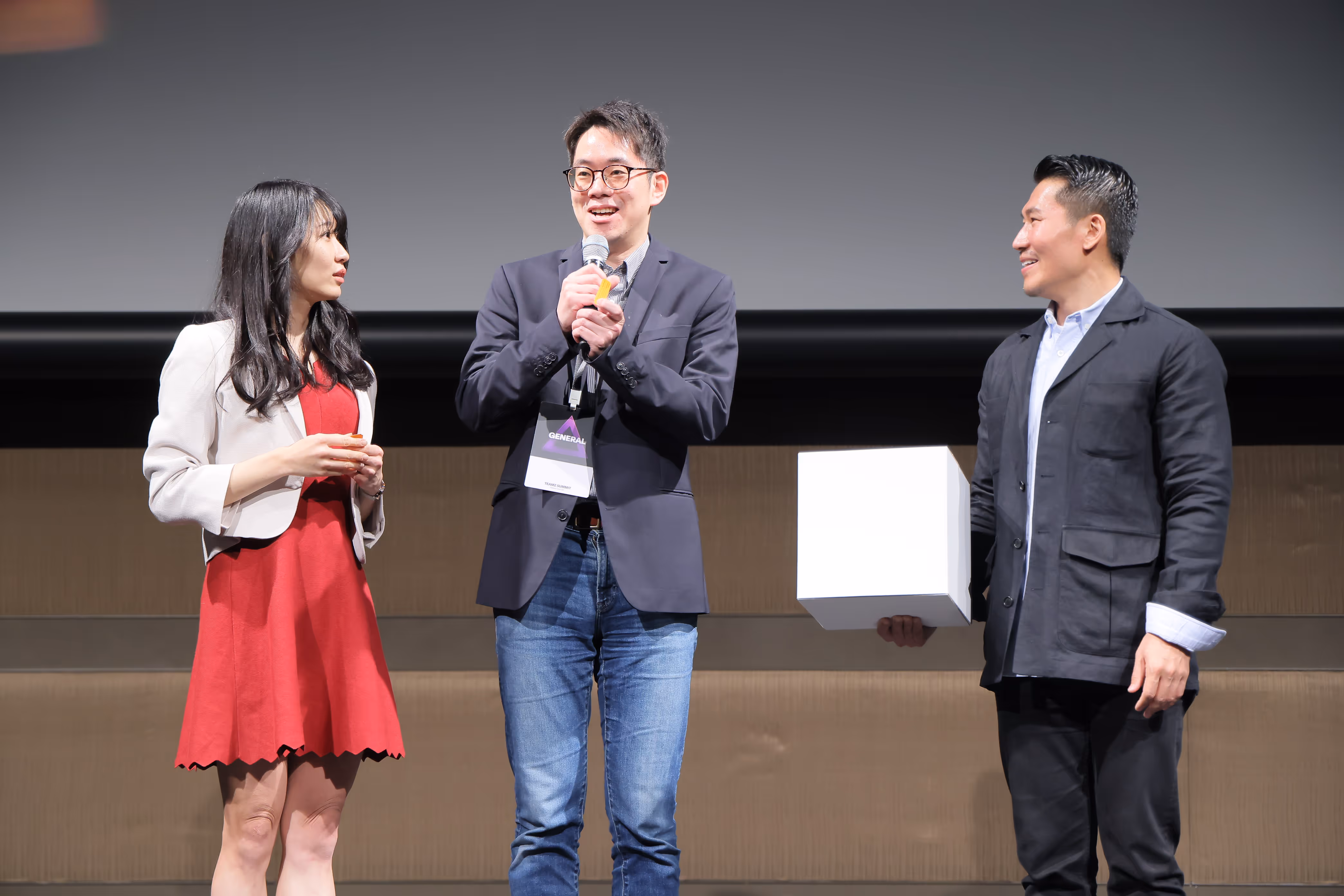 マイクを持って話している男性とその両脇で話を聞いている男女の三人の人々。