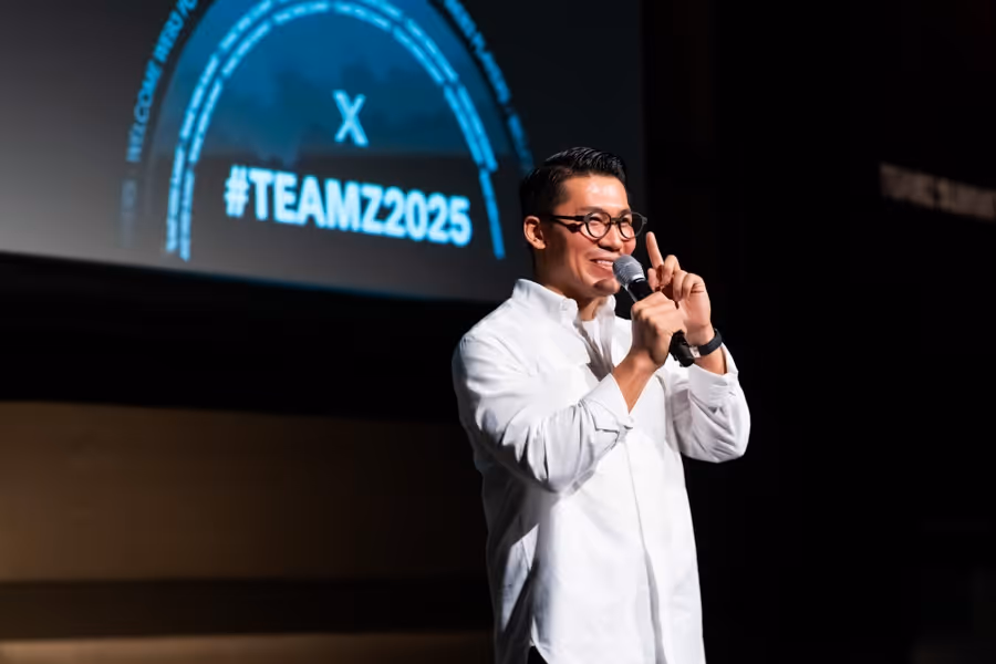 白いシャツを着た男性がマイクを持ち、指を立てて話している。