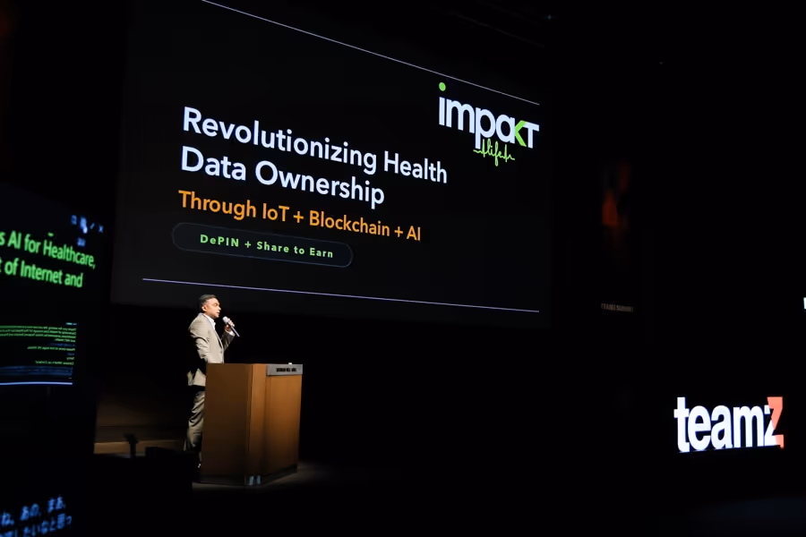 スーツを着た男性がステージでマイクを持ち、背後の大画面に「Revolutionizing Health Data Ownership Through IoT + Blockchain + AI」と表示されている。