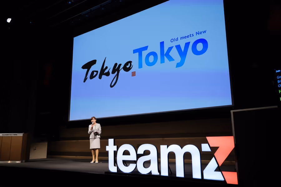 スーツを着た女性が「teamZ」と大きく表示されたステージで、『Tokyo Tokyo Old meets New』の背景スクリーンの前で講演している。