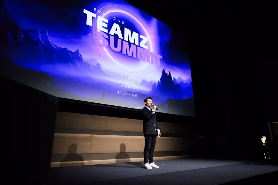 ステージでマイクを持ち発表している男性、背景に青紫色のTEAMZ SUMMIT 2026の大画面が映っている